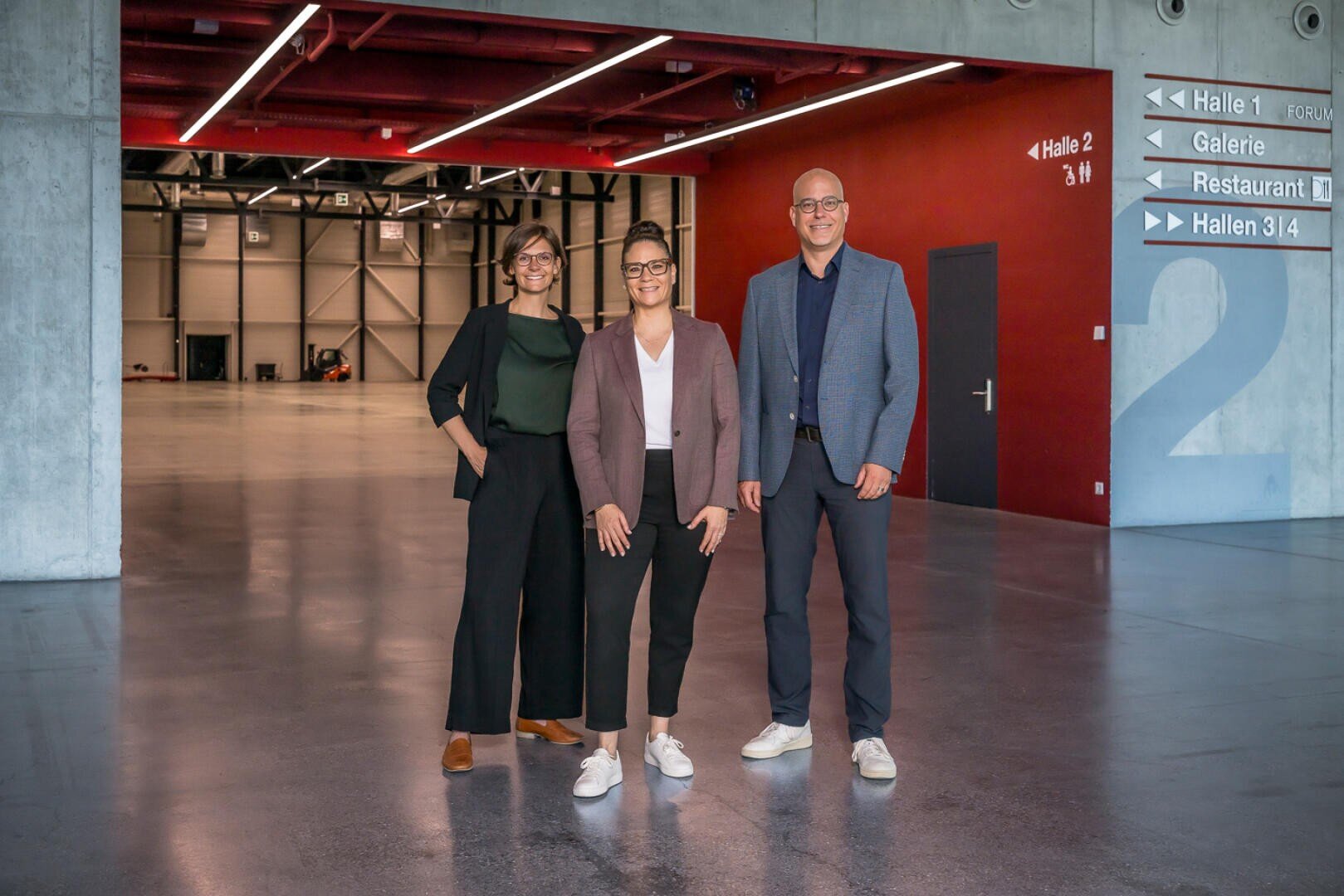 Drei Personen stehen lächelnd in einem modernen, geräumigen Gebäude mit roten und grauen Wänden, Betonböden und Wegweisern. Die beiden Frauen und der Mann sind im Business-Casual-Stil gekleidet.