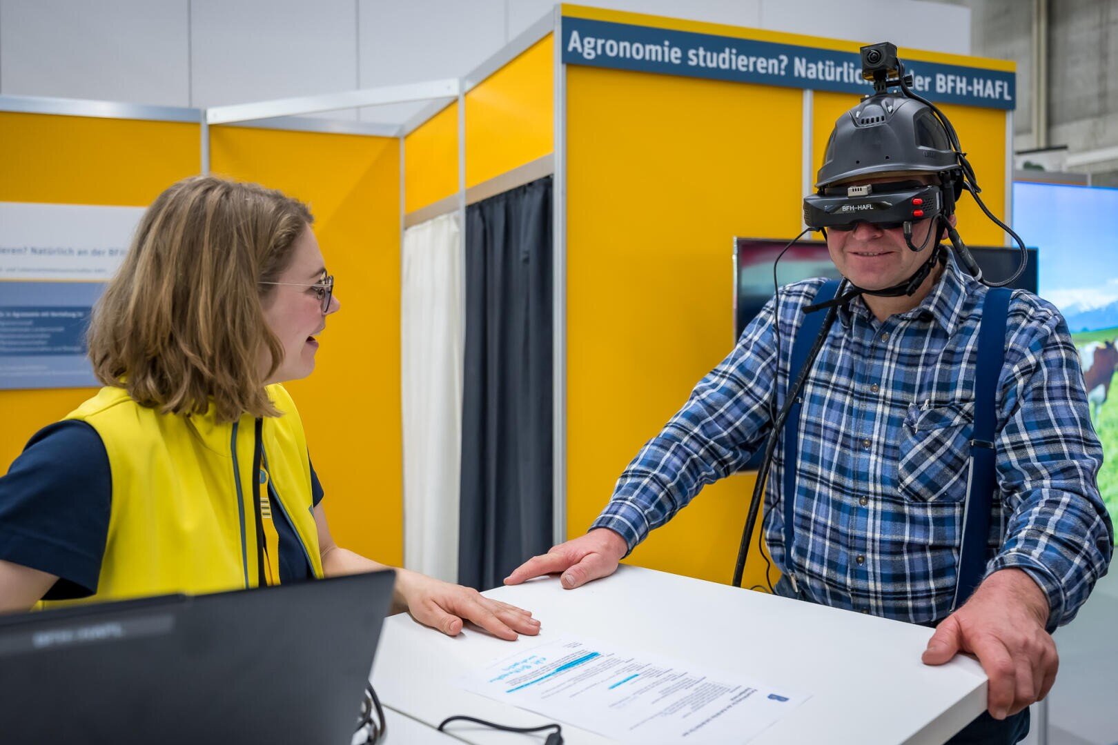 Eine Frau und ein Mann interagieren an einem Stand. Der Mann trägt einen Helm mit VR-Brille und Kameraausrüstung, während die Frau ihn anlächelt. Der Stand hat gelbe Wände und Informationsdisplays.