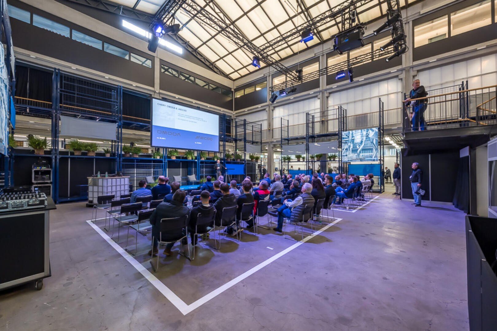 Eine Gruppe von Menschen, die in Reihen sitzen, hört einer Präsentation in einem großen, industriell anmutenden Raum mit hohen Decken, Metallgeländern und Projektionsflächen, auf denen Dias gezeigt werden, zu.