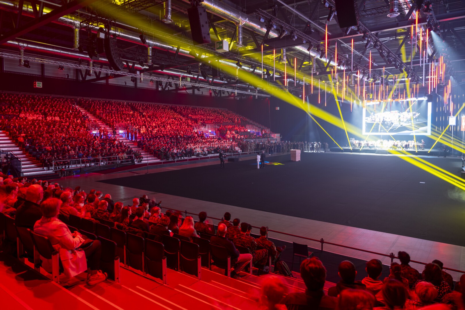 Eine große Indoor-Arena mit sitzenden Zuschauern, beleuchtet von roten und gelben Bühnenlichtern, gegenüber einer Bühne mit einem großen Bildschirm mit Grafiken. Die Atmosphäre ist lebhaft und energiegeladen.