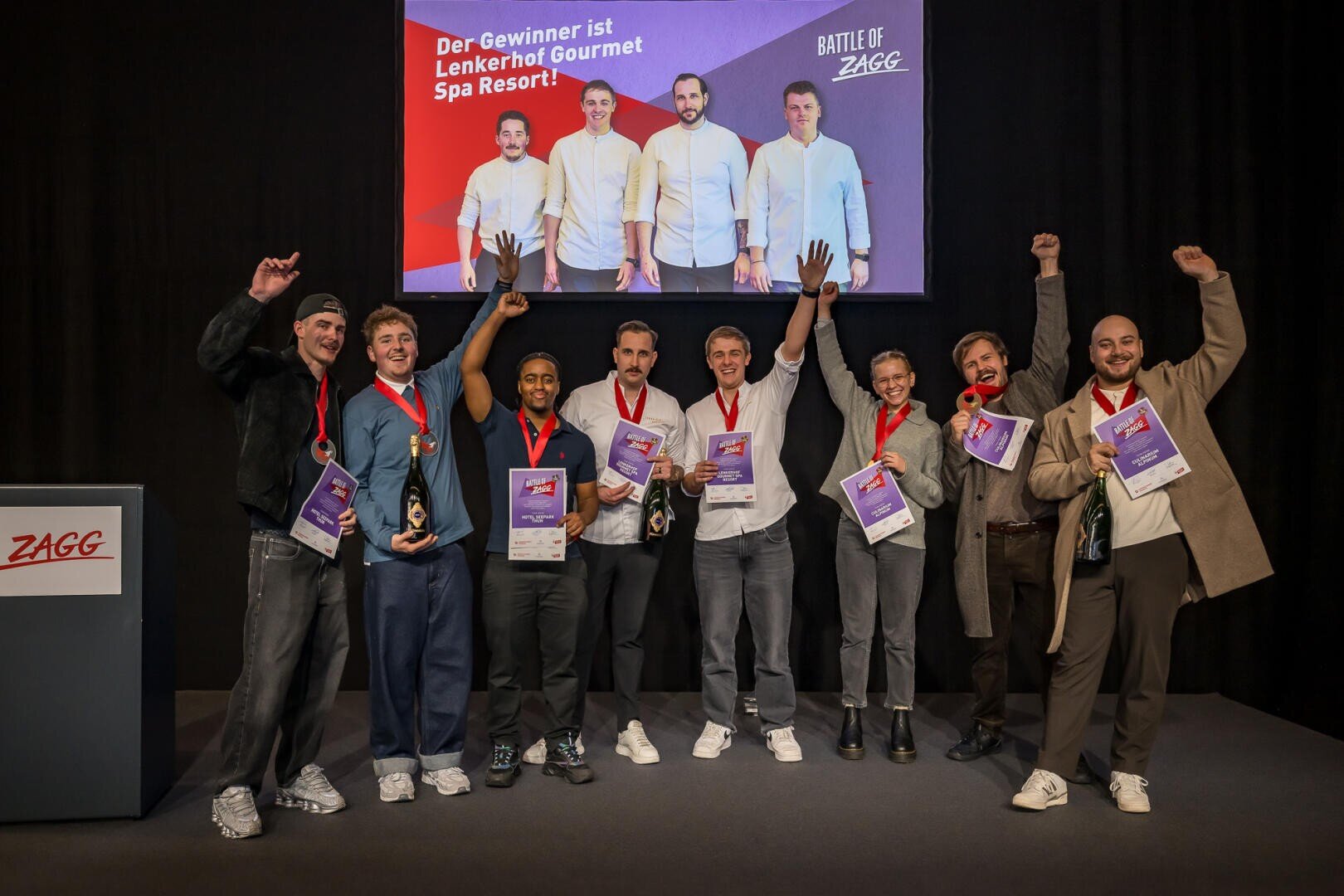 Eine Gruppe von acht Personen steht auf der Bühne und feiert mit Medaillen und Urkunden vor einer Leinwand, auf der der Text Der Gewinner ist das Lenkerhof Gourmet Spa Resort! und Battle of ZAGG zu lesen ist.
