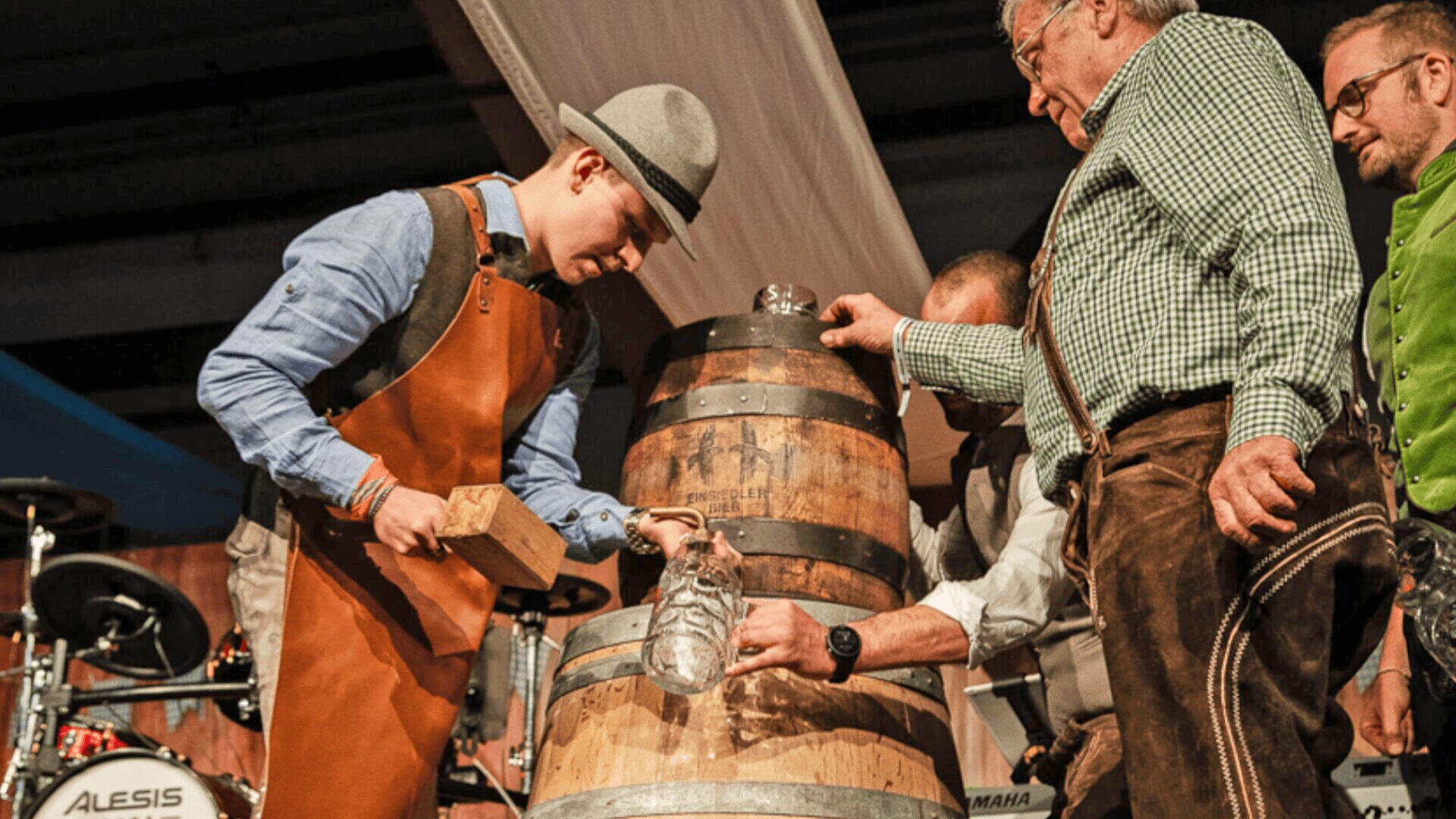Ein Mann mit Schürze und Hut zapft mit einem Hammer ein hölzernes Bierfass an. Ältere Männer in traditioneller Kleidung helfen ihm dabei, was wie ein Fest oder eine Bierveranstaltung aussieht.