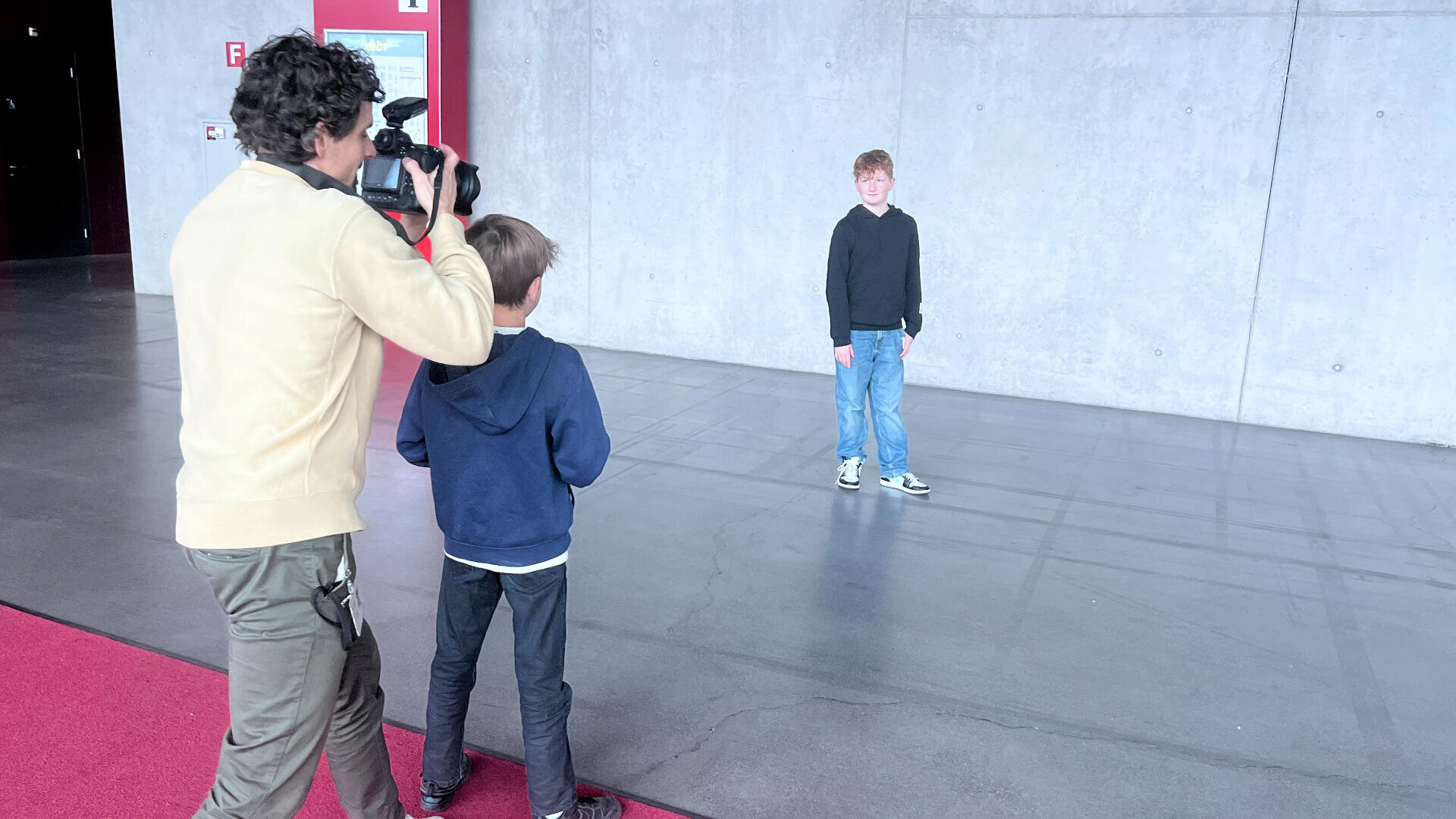 Ein Kameramann filmt einen kleinen Jungen, der vor einer großen grauen Betonwand steht, während ein anderes Kind zusieht. Die Szenerie scheint ein öffentlicher Innenraum zu sein, an dessen Seite ein roter Teppich liegt.