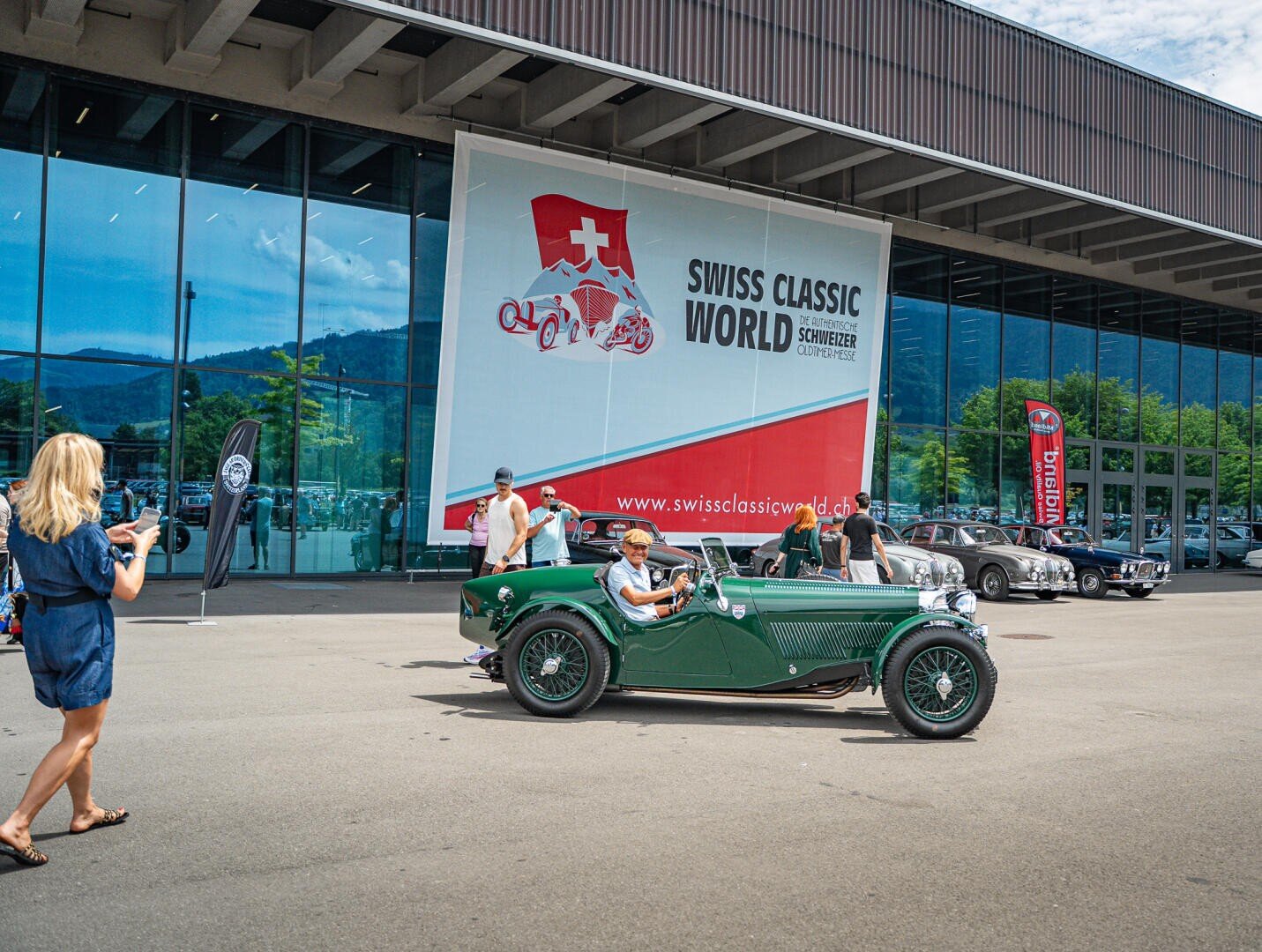 Ein grüner Oldtimer fährt am Veranstaltungsgebäude der „Swiss Classic World“ vorbei, während Menschen Fotos machen und weitere Oldtimer in der Nähe geparkt sind.