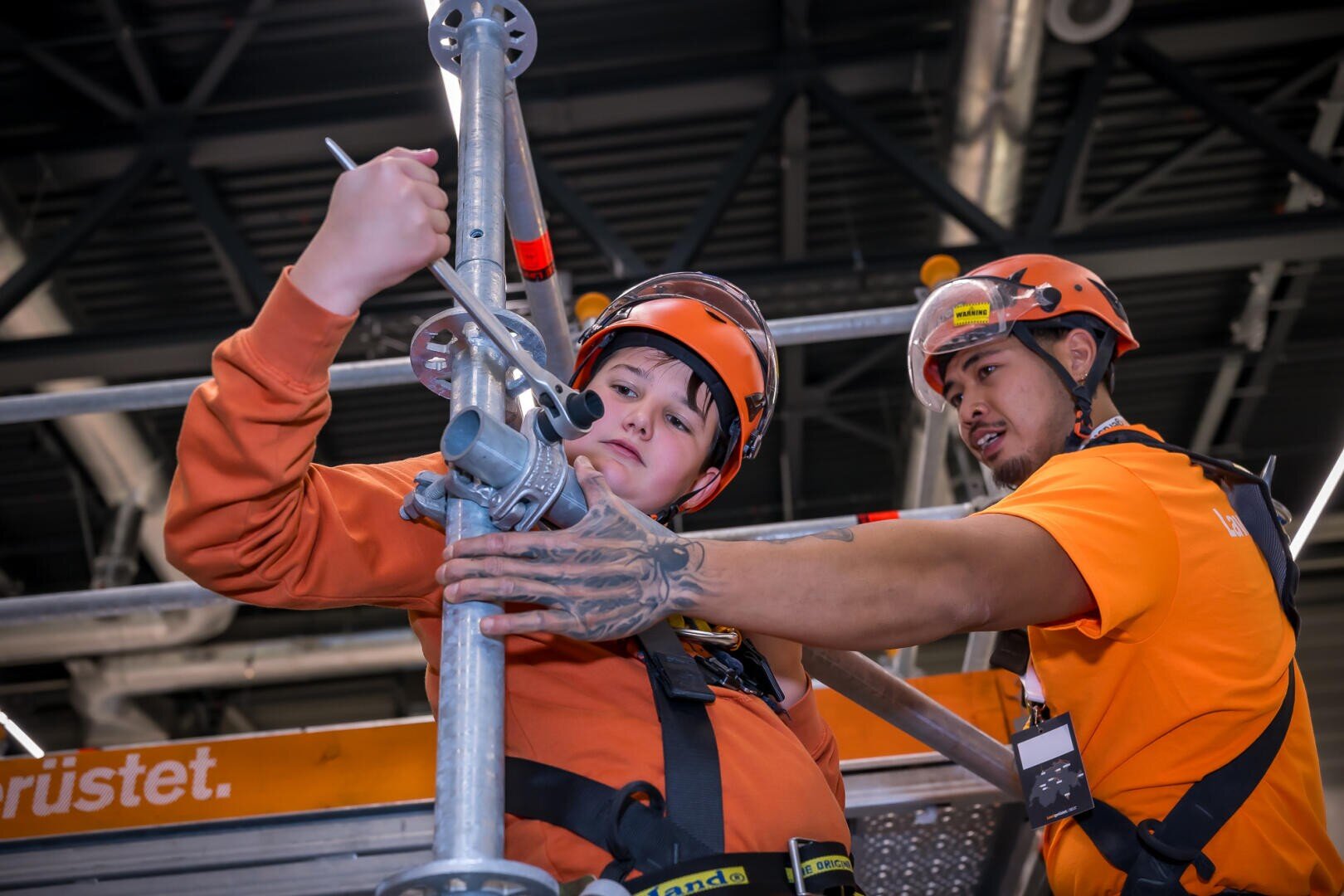 Zwei Bauarbeiter in orangefarbener Schutzkleidung und mit Helmen bauen in einem Gebäude ein Gerüst auf. Der eine justiert eine Metallstange, während der andere hilft, beide konzentrieren sich auf ihre Aufgabe. Im Hintergrund sind Industrieträger und Ausrüstung zu sehen.