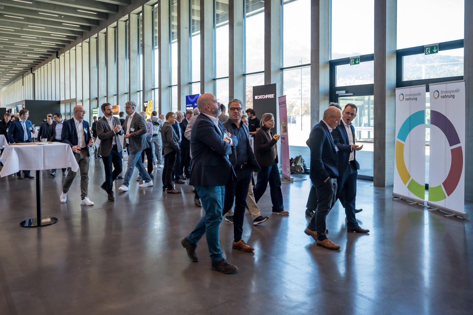 Eine Gruppe von Personen in Geschäftskleidung geht durch einen modernen, sonnendurchfluteten Konferenzsaal mit großen Fenstern, Ausstellungsbannern und runden Tischen im Hintergrund.
