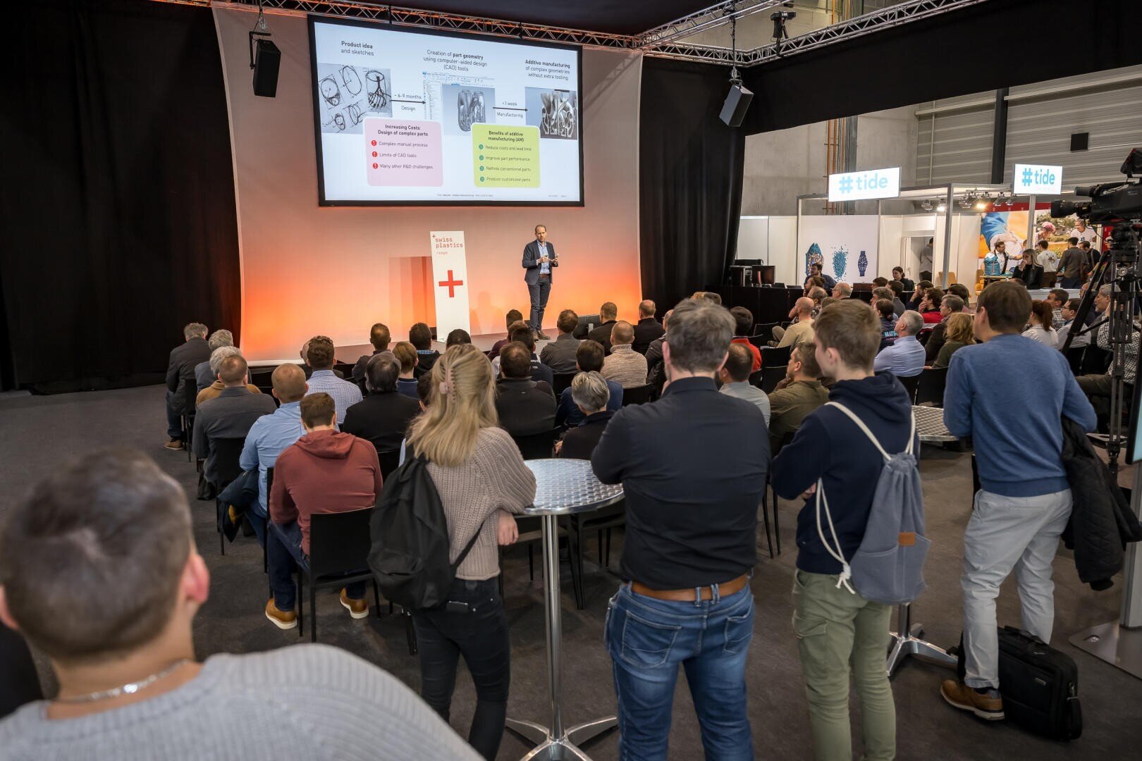 Ein Redner hält einen Vortrag vor einem sitzenden Publikum in einem großen Konferenzraum. Hinter ihm werden Folien auf eine Leinwand projiziert, und im Hintergrund sind Messestände zu sehen.