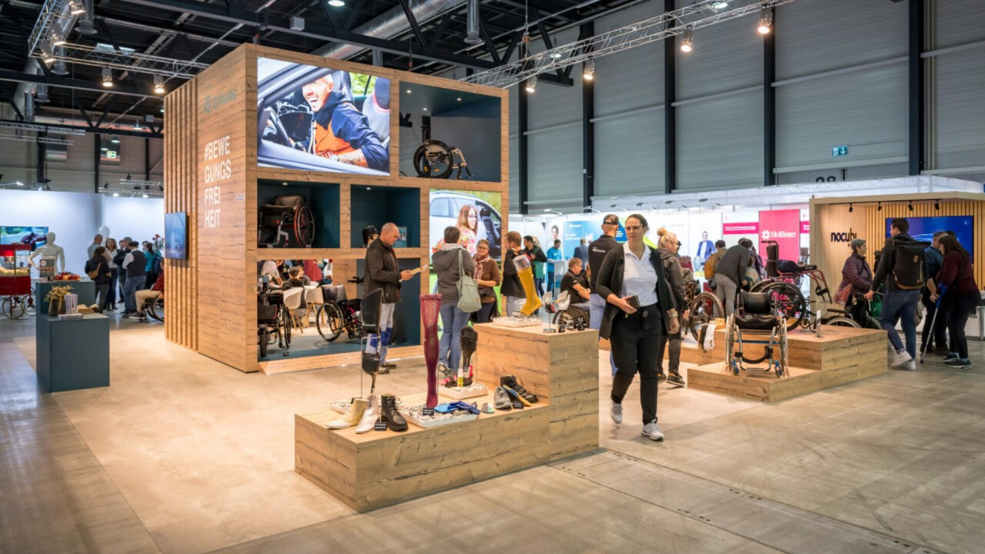 In einer geschäftigen Kongresshalle erkunden Menschen einen großen Holzstand, an dem Rollstühle und Prothesen ausgestellt sind. Puppen präsentieren Mobilitätshilfen und auf einem großen Bildschirm werden entsprechende Bilder gezeigt.