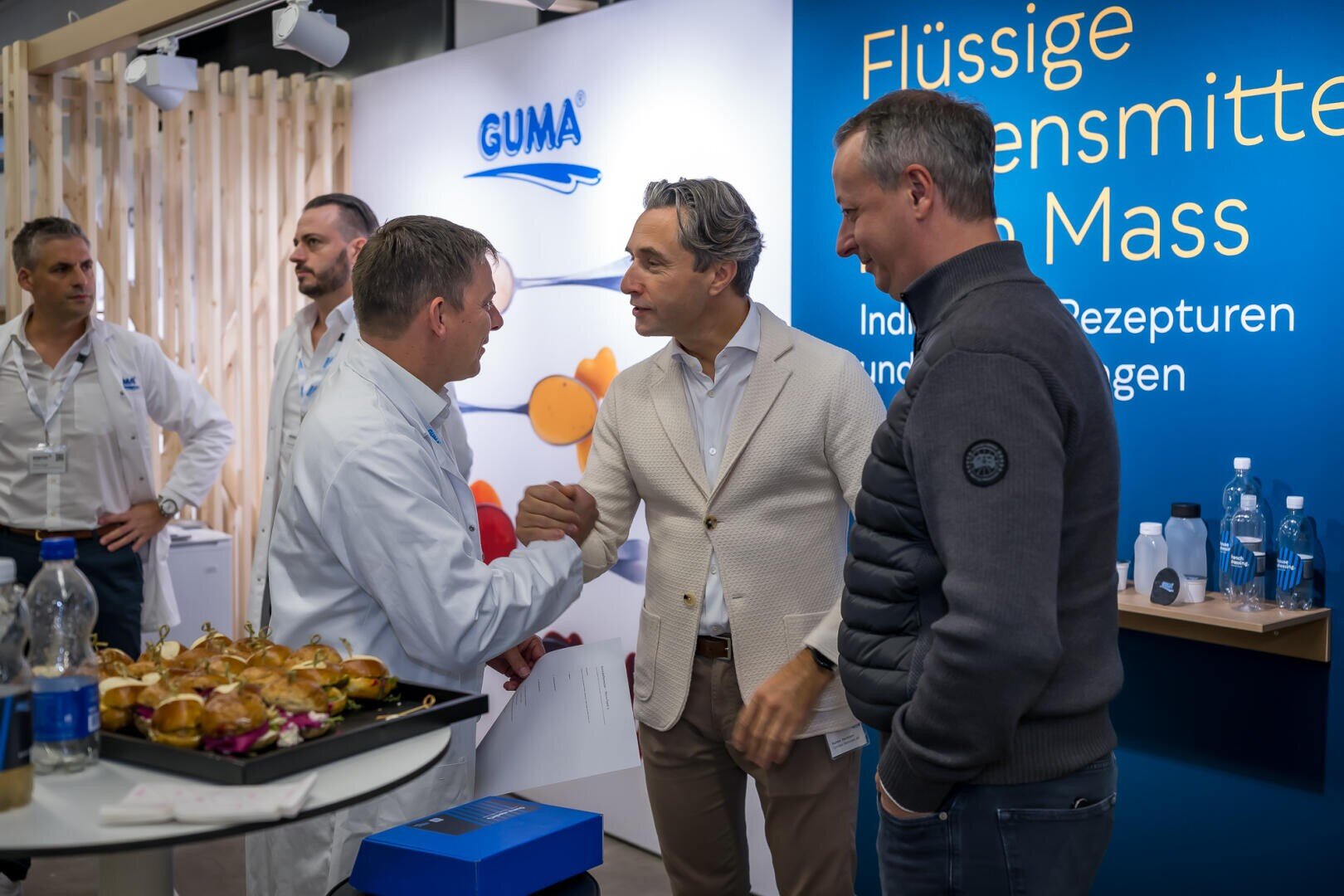 Vor einem Ausstellungsstand der Firma Guma schüttelt ein Mann in einem weißen Laborkittel einem Mann in einem beigen Blazer die Hand, während ein dritter Mann in einer dunklen Jacke zusieht.