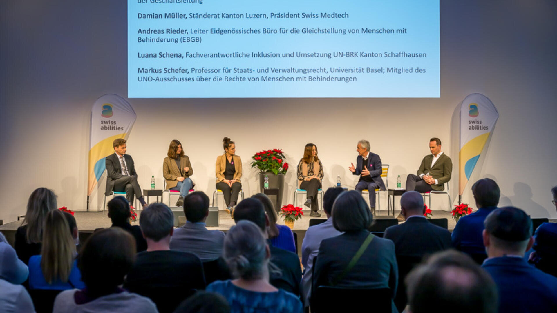 Ein fünfköpfiges Gremium sitzt mit Mikrofonen auf der Bühne und blickt auf das Publikum. Dahinter befindet sich ein großer Bildschirm mit Texten und Namen; Blumenarrangements schmücken die Bühne. Auf beiden Seiten sind die Logos von Swiss Abilities zu sehen.