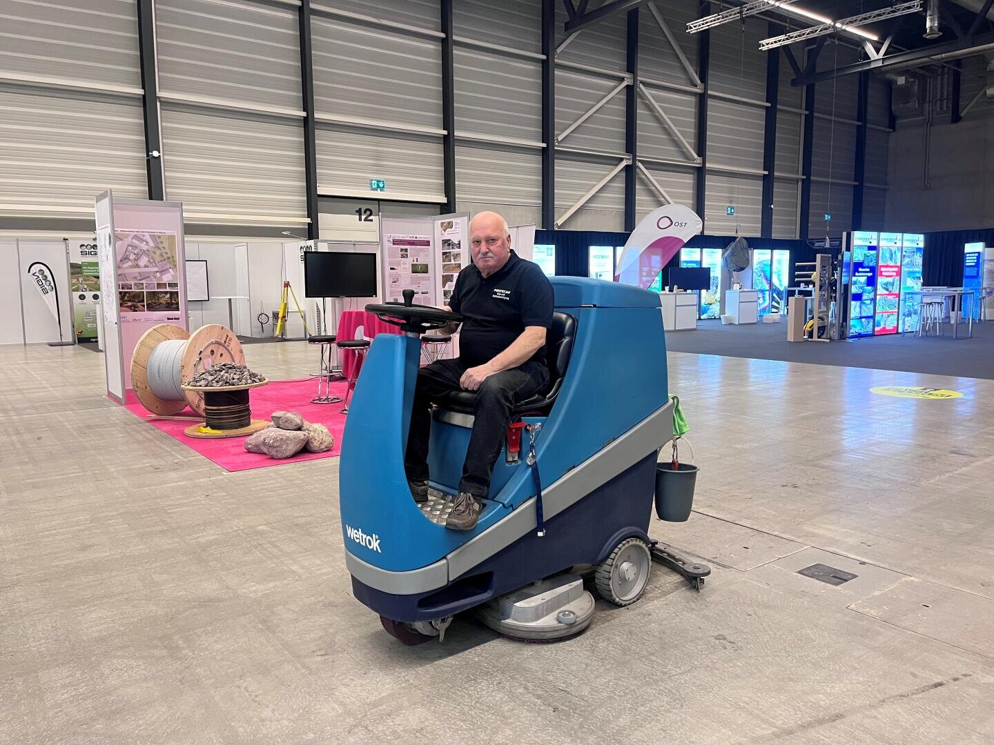 Ein Mann fährt auf einer großen blauen Bodenreinigungsmaschine in einer geräumigen Ausstellungshalle mit Ständen, Displays und heller Beleuchtung im Hintergrund.