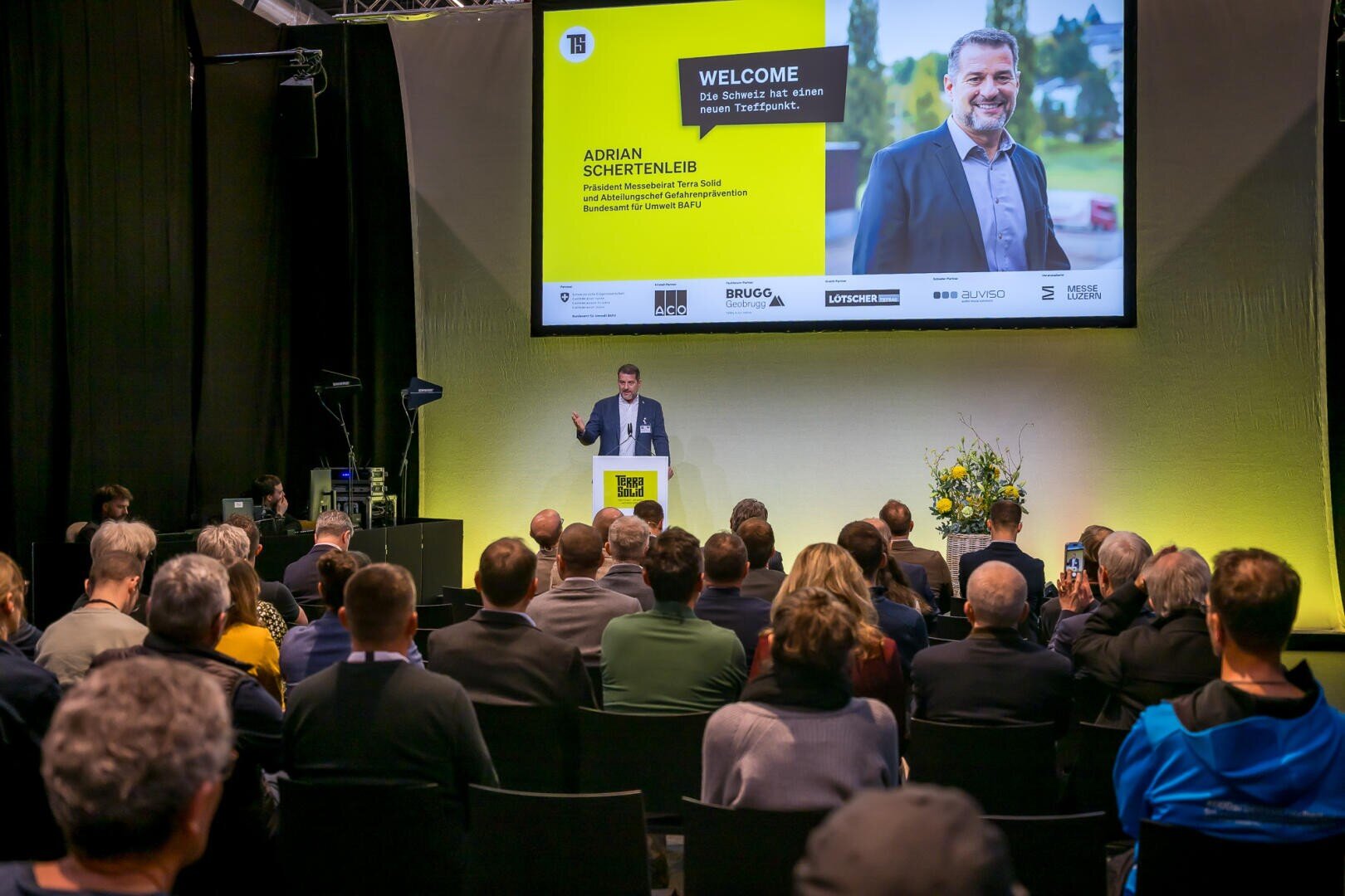 Ein Mann steht an einem Podium und spricht zu einem Publikum in einem Konferenzraum. Hinter ihm befindet sich ein großer Bildschirm, auf dem sein Foto, sein Name (Adrian Schertenleib) und eine Begrüßungsbotschaft angezeigt werden. Das Publikum sitzt und hört aufmerksam zu.