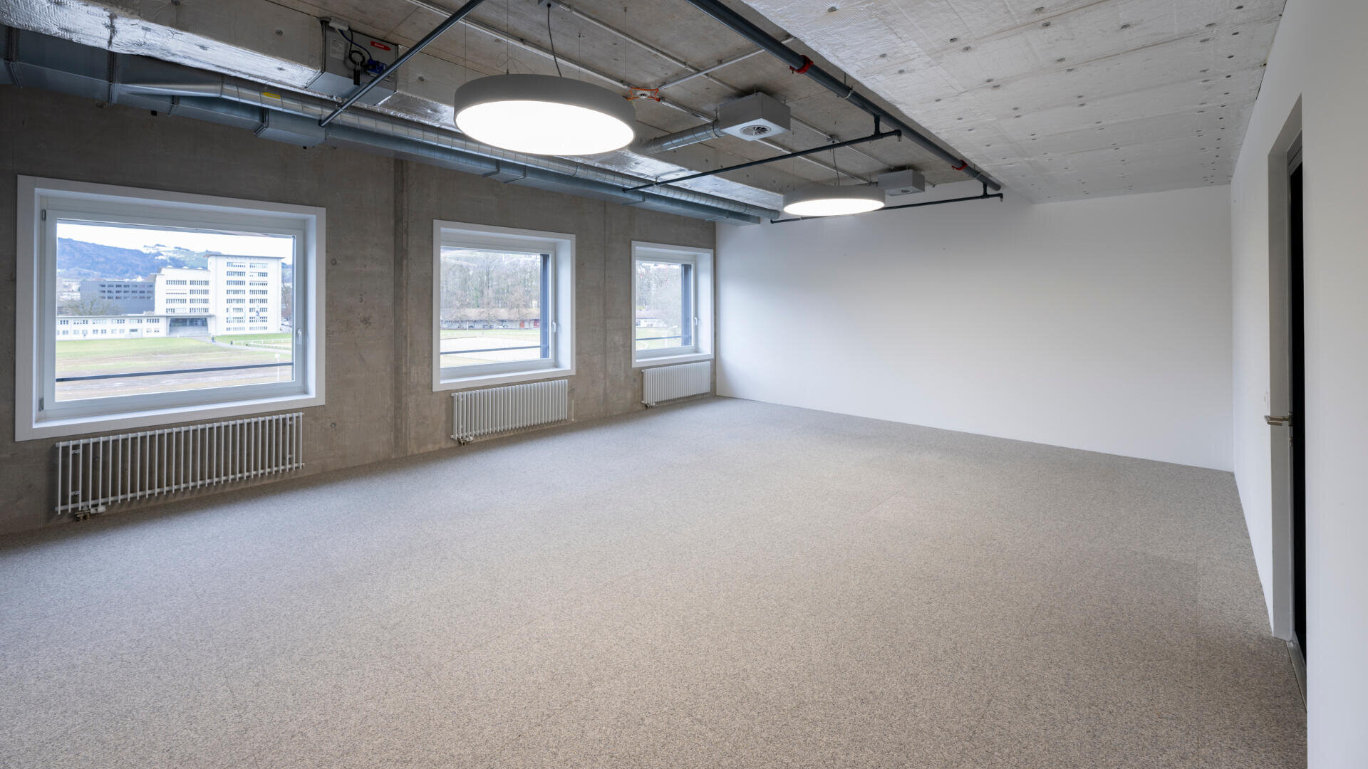 Ein moderner, leerer Büroraum mit grauem Teppichboden, drei großen Fenstern, Sichtbetondecke, runden Deckenleuchten und einer weißen Wand. Draußen sind Gebäude und Grünflächen sichtbar.
