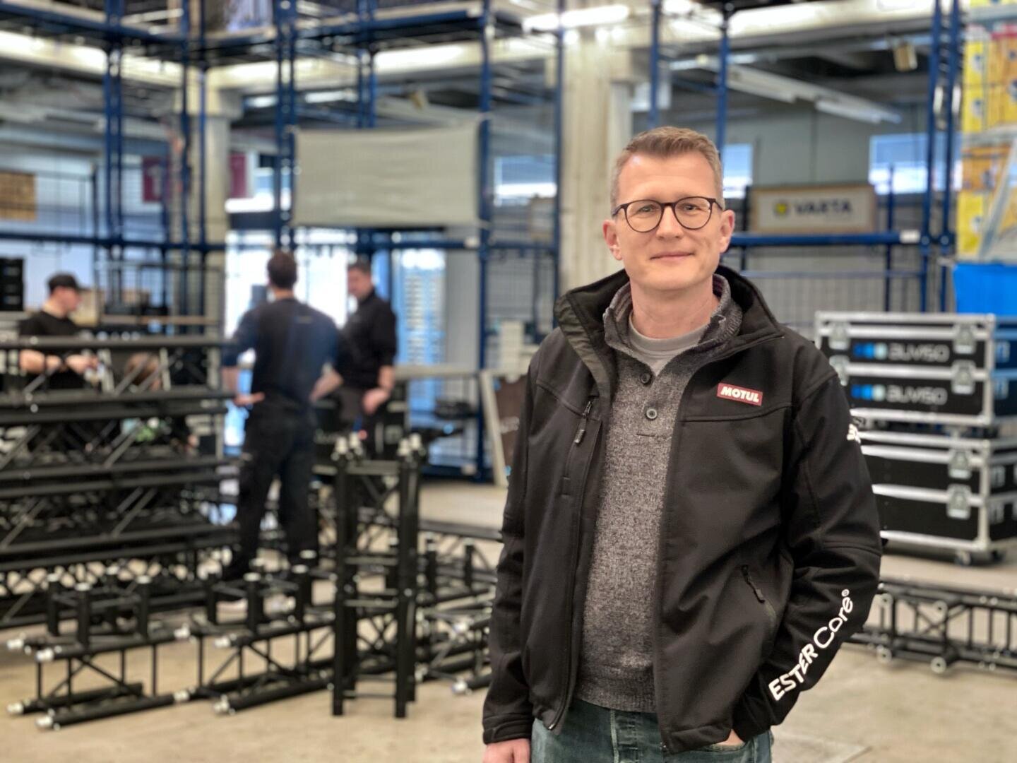 Ein Mann mit Brille und schwarzer Jacke steht lächelnd in einem Industrielager mit Metallstrukturen und Kisten. Im Hintergrund sind mehrere Personen dabei, Geräte zu montieren.