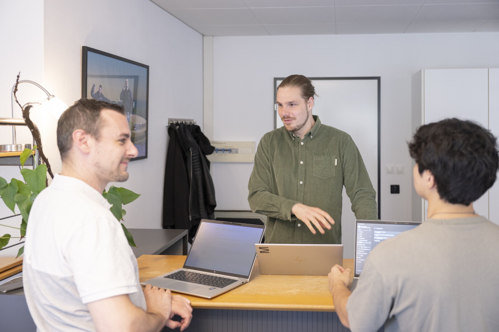 Drei Personen stehen mit Laptops an einem Schreibtisch und unterhalten sich. Eine Person steht hinter dem Schreibtisch, die beiden anderen stehen davor und blicken ihn an. Die Szenerie wirkt wie ein Büro mit Pflanzen und Wanddekoration.
