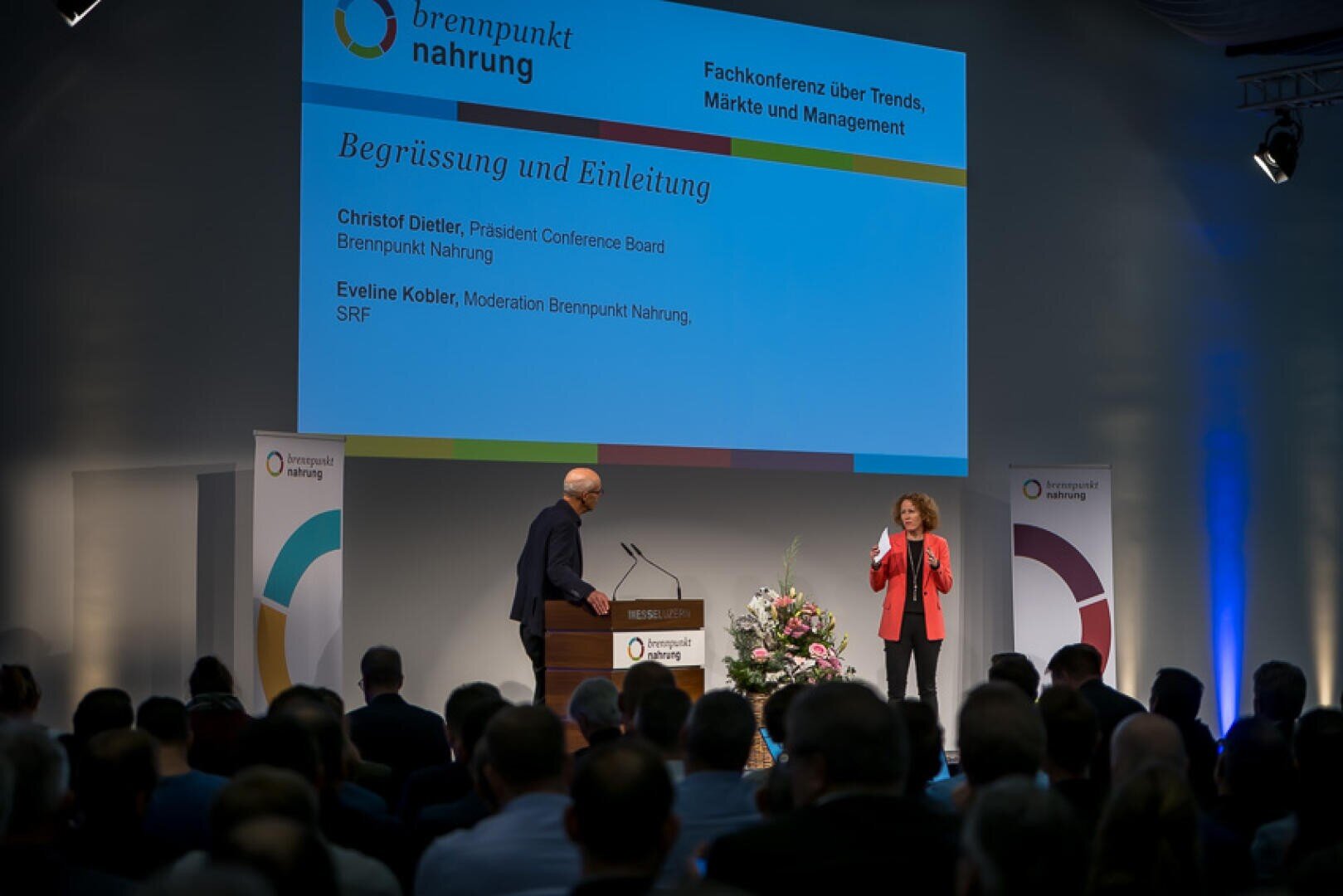 Ein Mann und eine Frau halten auf der Bühne einer Konferenz namens „brennpunkt nahrung“ einen Vortrag. Das Publikum sitzt, und auf einer großen Leinwand hinter ihnen werden die Namen der Redner sowie Details zur Veranstaltung auf Deutsch angezeigt.
