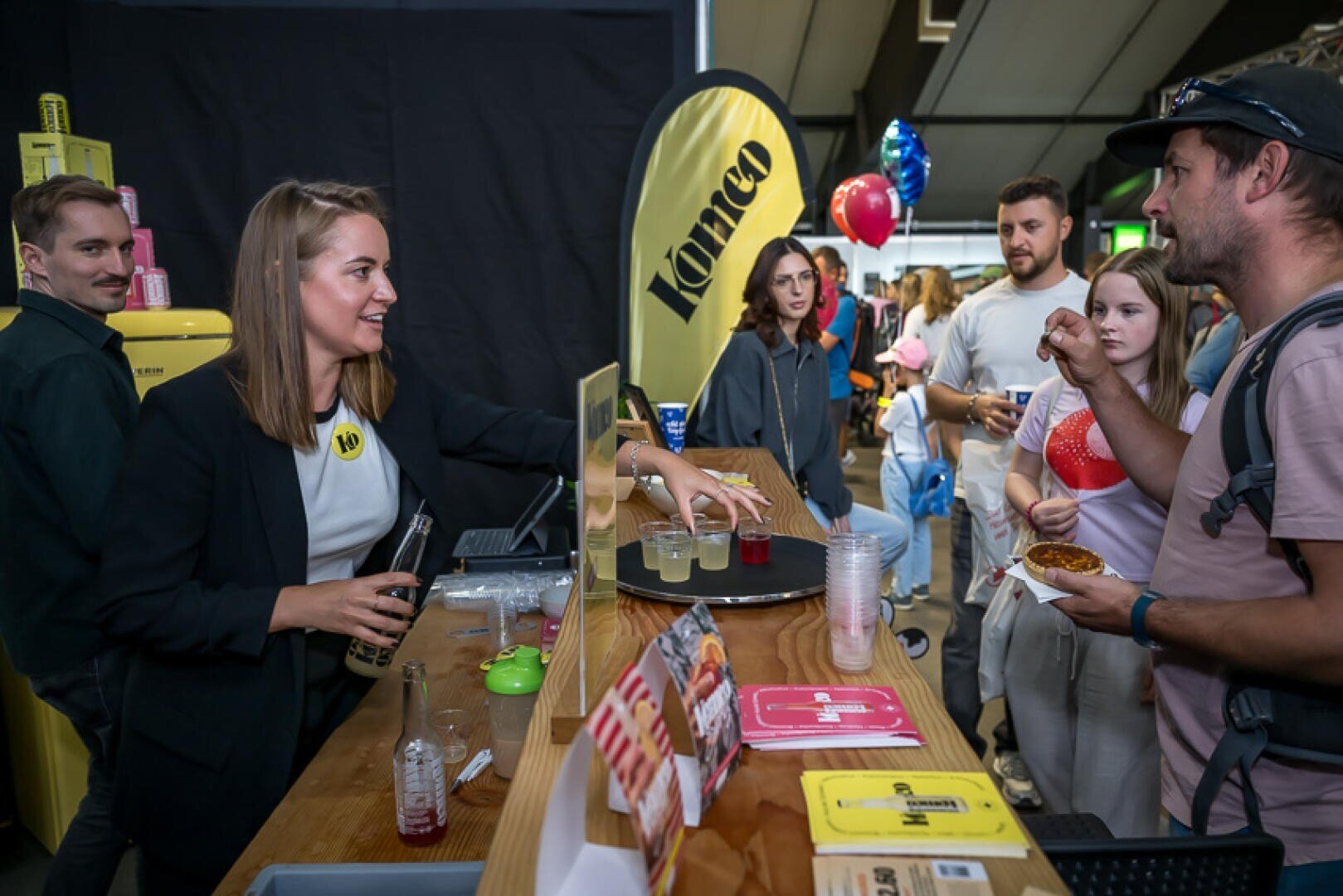 Eine Frau hinter der Theke eines Standes schenkt bei einer Veranstaltung in einem Saal einer Gruppe von Menschen Getränke ein. Auf der Theke stehen Becher, Flyer und Flaschen, und die Besucher stehen Schlange, wobei einige Essen und Getränke in den Händen halten.