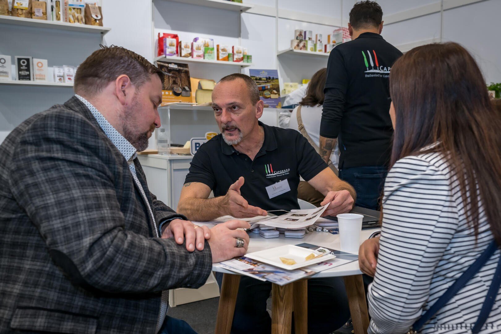 Ein Mann in einem Marken-Poloshirt gestikuliert, während er sich auf einer Messe mit zwei Personen über einen kleinen Tisch hinweg unterhält.