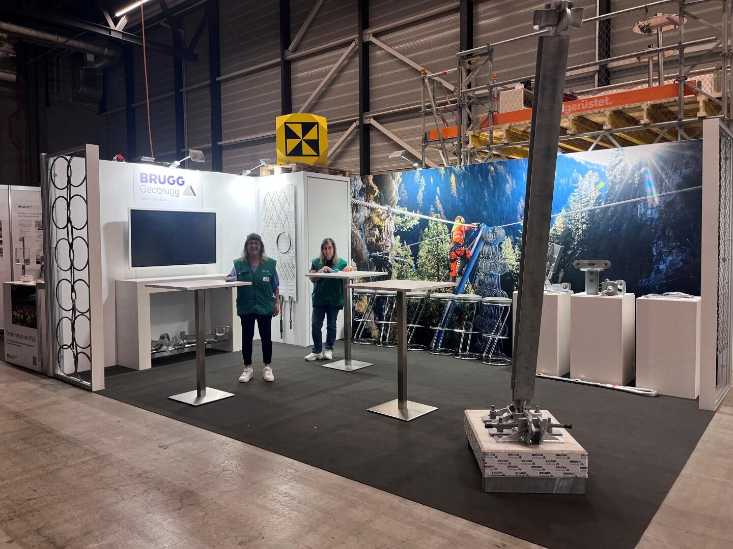 Zwei Personen stehen an einem Messestand von BRUGG Geobrugg mit Ausstellungstischen, Industrieausrüstungen und einem großen Foto einer Person, die an einem Bergsicherungssystem arbeitet. Der Stand befindet sich in einer großen Halle.