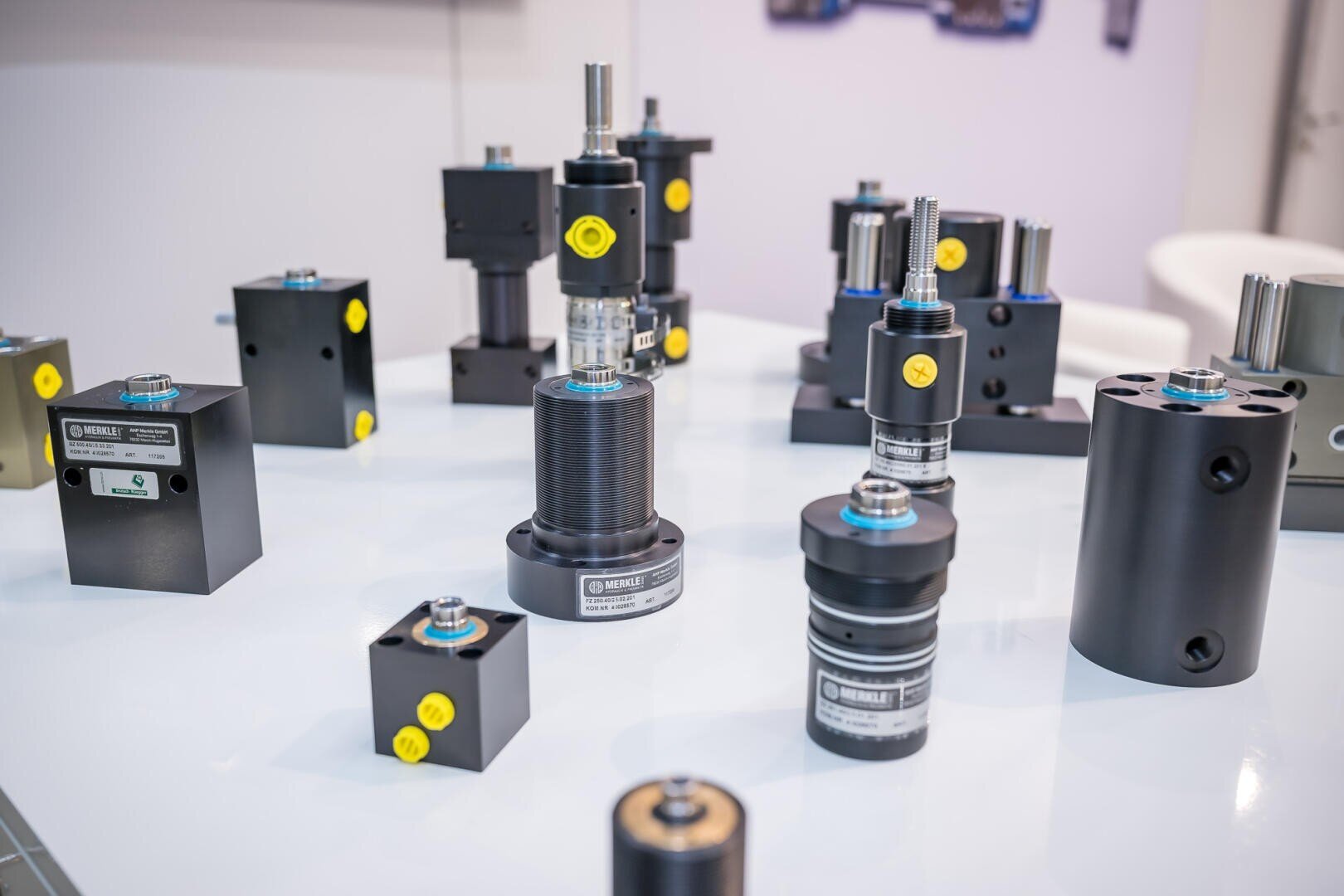 Eine Auslage mit verschiedenen industriellen Maschinenteilen, darunter zylindrische und rechteckige Metallteile mit Etiketten und gelben Kappen, die auf einem weißen Tisch angeordnet sind.