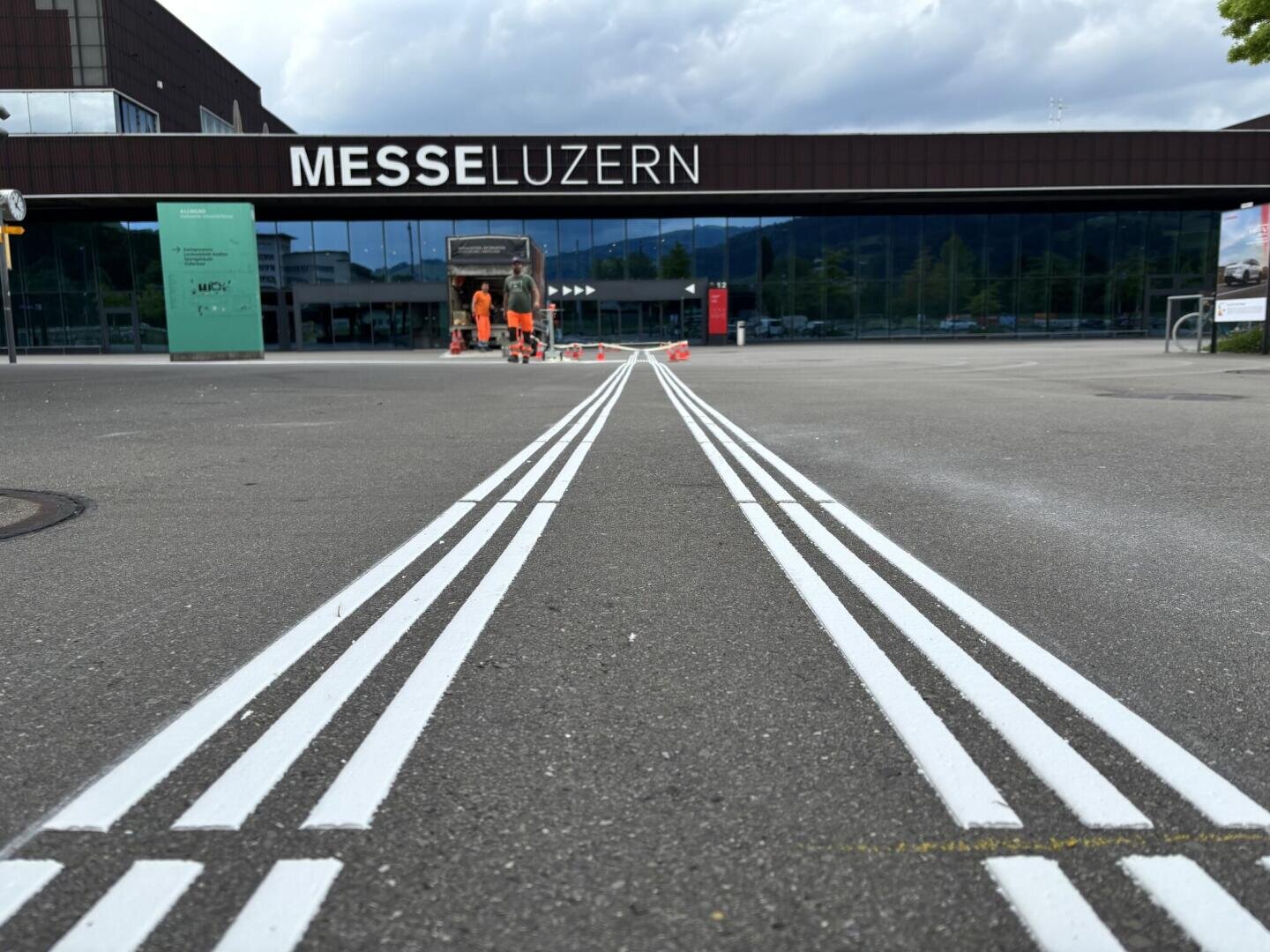 Frisch gestrichene weiße Fahrbahnmarkierungen führen zum Eingang der Messe Luzern. Im Hintergrund sind Arbeiter in orangefarbenen Uniformen und ein Fahrzeug zu sehen. Die Szene spielt im Freien, der Himmel ist bewölkt.