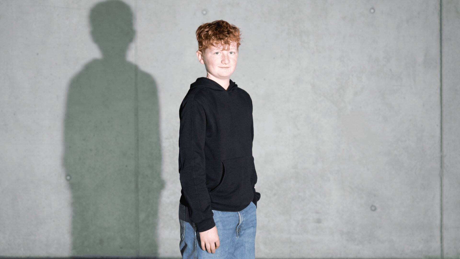 Eine Person mit lockigem rotem Haar, die einen schwarzen Kapuzenpullover und blaue Jeans trägt, steht vor einer hellgrauen Betonwand und wirft einen deutlichen Schatten nach links.