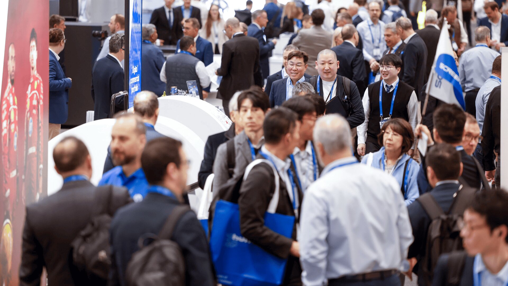 Eine überfüllte Messe- oder Ausstellungshalle ist voller Menschen, die spazieren gehen, sich unterhalten und Business-Kleidung und blaue Schlüsselbänder tragen. Manche tragen blaue Tragetaschen. Stände und Banner säumen den belebten Gehweg.