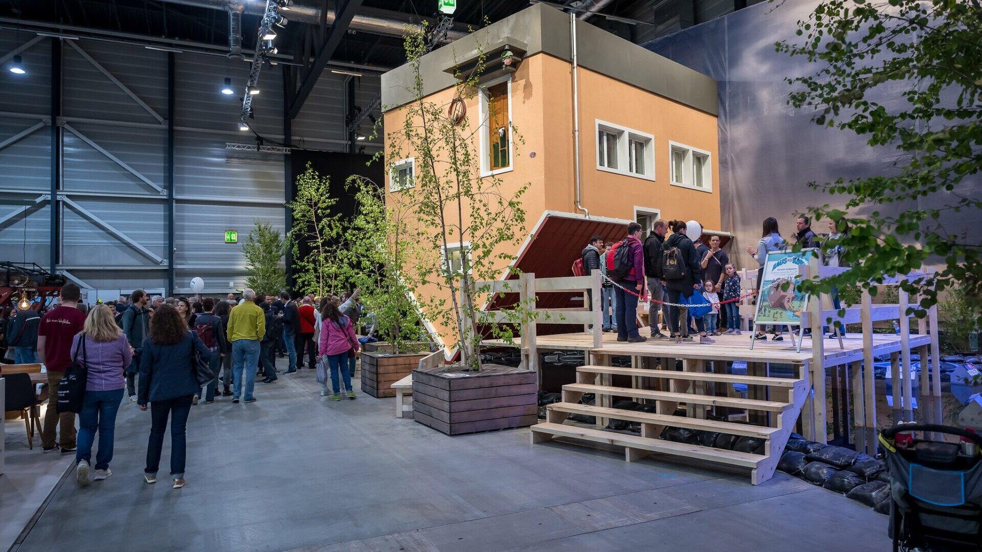 Auf einer Ausstellung oder Messe versammeln sich die Menschen um ein Modellhaus; einige stehen Schlange, um hineinzukommen, andere gehen umher, und überall sind Bäume und Pflanzen aufgestellt.