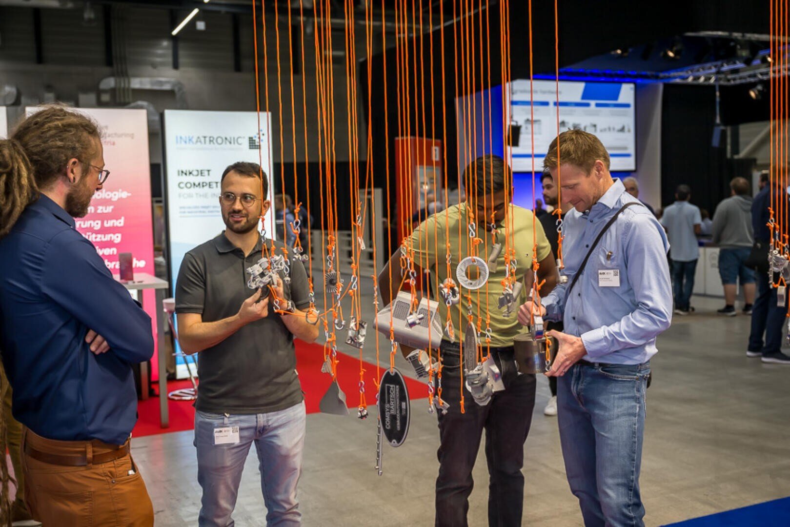 Vier Männer stehen um eine Auslage mit hängenden mechanischen Teilen in einer Technologieausstellung. Sie unterhalten sich und begutachten die Gegenstände, während im Hintergrund Informationsbanner und Bildschirme zu sehen sind.