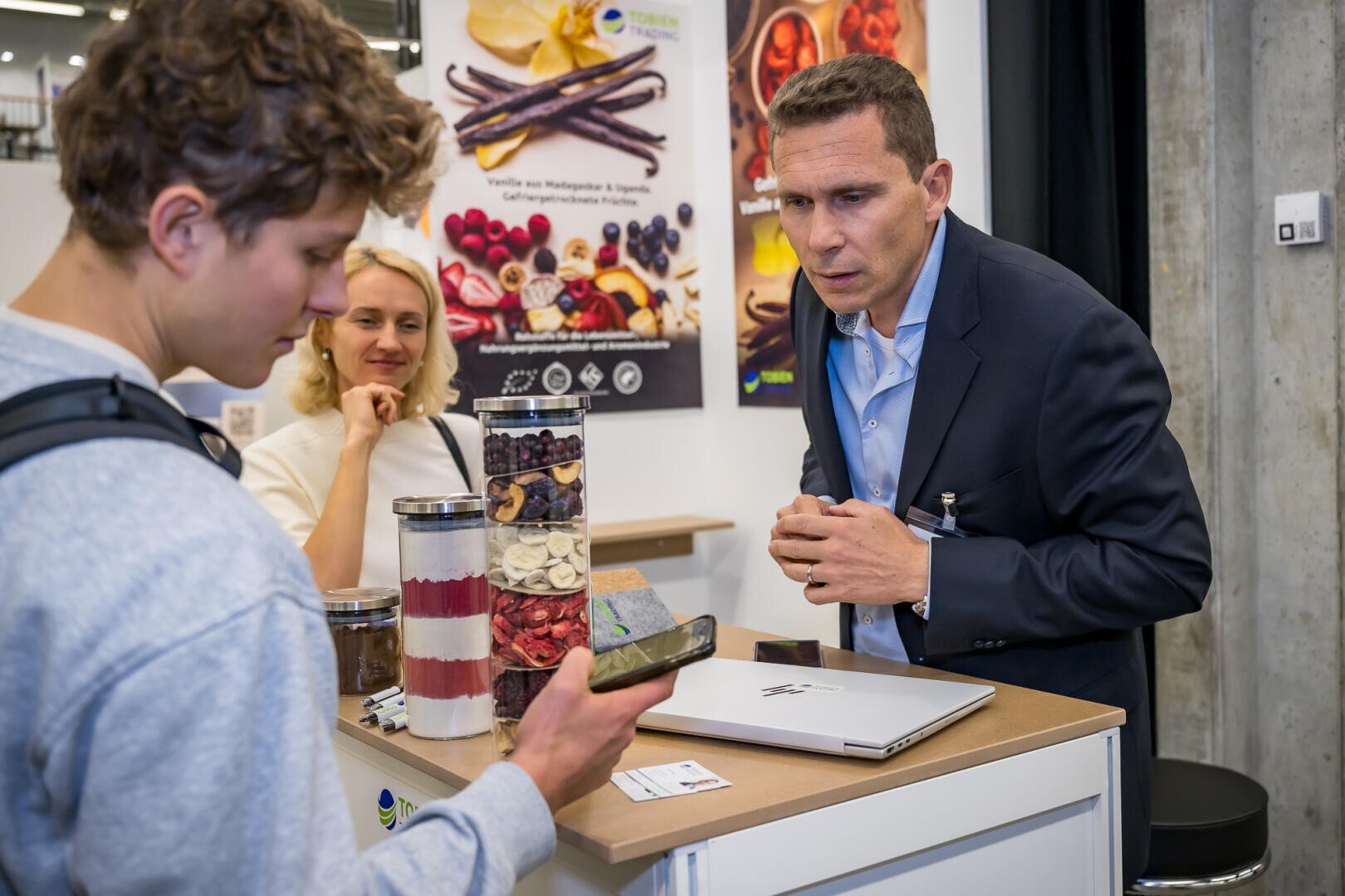 Ein Mann im Anzug unterhält sich mit einem Kunden an einem Messestand, auf dem Gläser mit getrockneten Zutaten ausgestellt sind.