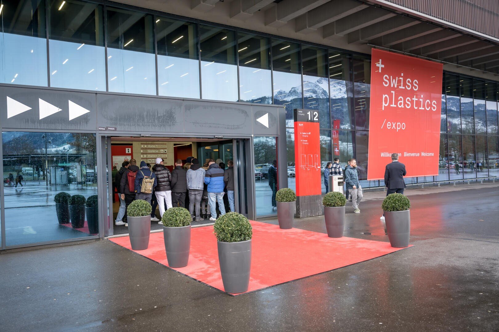 Eine Gruppe von Personen betritt ein modernes Gebäude mit großen Glasfenstern und einem roten Teppich am Eingang. Auf einem Schild steht "Swiss Plastics Expo". Im Hintergrund sind schneebedeckte Berge zu sehen.