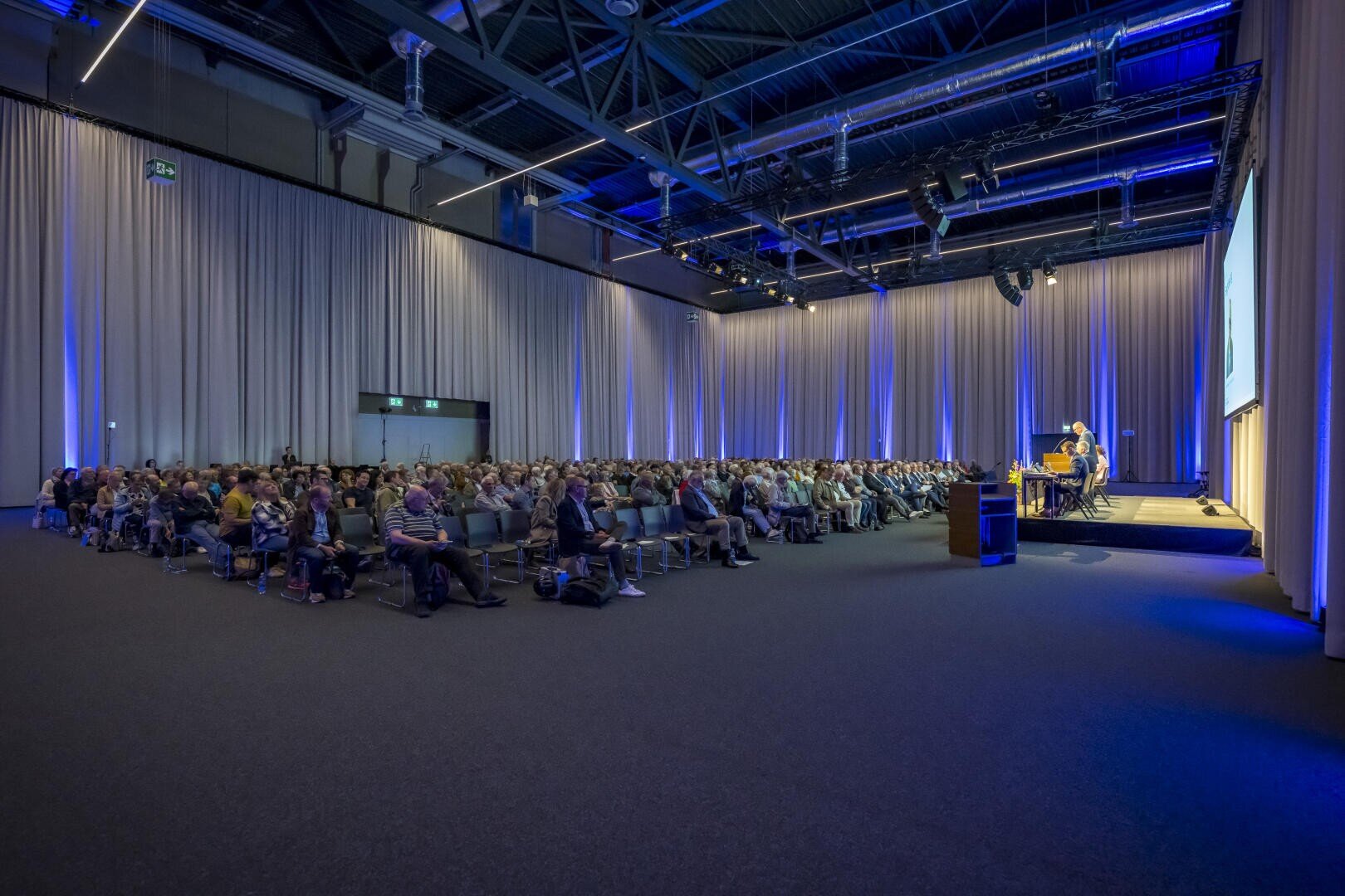 Ein großes Publikum sitzt in Reihen vor einer Bühne, auf der ein Redner bei blauer Beleuchtung in einem großen Konferenzsaal mit hohen Decken und weißen Vorhängen spricht.