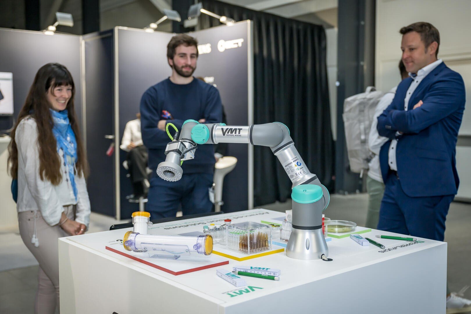 Drei Personen stehen bei einer Indoor-Veranstaltung oder -Ausstellung um einen Demonstrationstisch mit einem Roboterarm und verschiedenen wissenschaftlichen Gegenständen. Die Gruppe wirkt engagiert und interessiert an der Ausstellung.