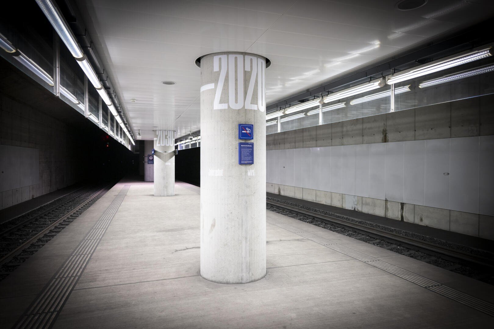 Ein leerer, moderner U-Bahn-Bahnhof mit Betonsäulen mit der Aufschrift „2020“, zwei Gleisen und heller Deckenbeleuchtung. Der Bahnsteig ist sauber und menschenleer.