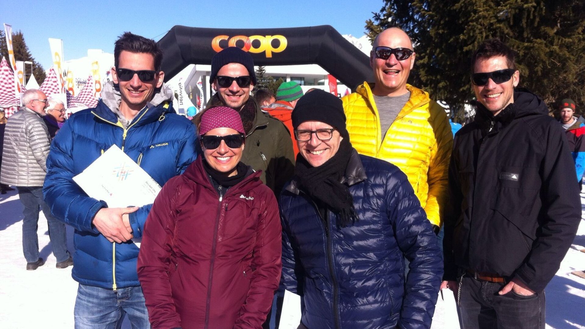 Eine Gruppe von sechs lächelnden Erwachsenen mit Sonnenbrillen und Winterjacken posiert an einem sonnigen, verschneiten Tag im Freien, mit anderen Menschen, Flaggen und einem aufblasbaren Bogen mit Coop-Logo im Hintergrund.
