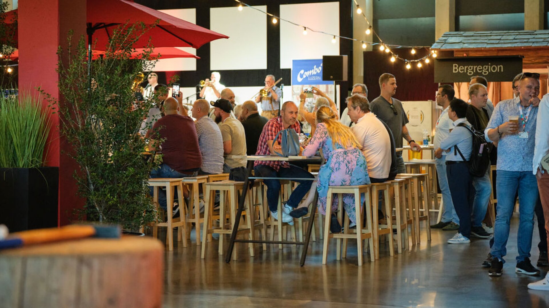 Bei einer Indoor-Veranstaltung mit Lichterketten, Pflanzen und Essensständen sitzen und stehen Menschen um Holztische und -hocker herum. Die Atmosphäre ist lebhaft und gesellig, die Gruppen unterhalten sich und essen.