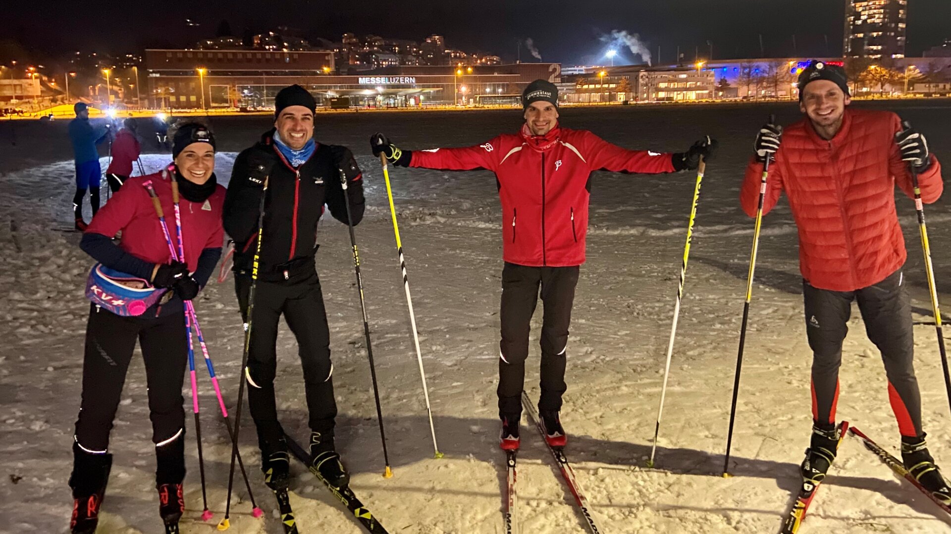 Vier Personen in Wintersportkleidung stehen nachts auf einem verschneiten Feld. Sie halten Skistöcke in der Hand und sind auf Langlaufskiern unterwegs. Im Hintergrund sind beleuchtete Stadtgebäude zu sehen. Sie lächeln und posieren für das Foto.