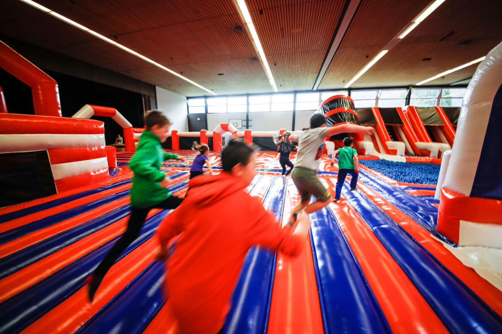 Die Kinder rennen und spielen auf einer großen aufblasbaren Hindernisbahn mit roten, blauen und weißen Streifen in einer geräumigen Halle mit hohen Decken und natürlichem Licht.