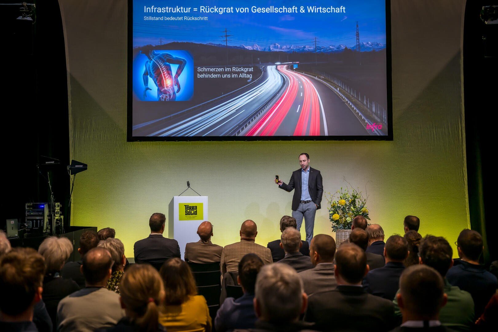 Ein Mann im Anzug hält auf der Bühne einen Vortrag vor einem Publikum. Hinter ihm ist ein großer Bildschirm in deutscher Sprache zum Thema Infrastruktur zu sehen, der eine nächtliche Autobahn mit Lichtspuren und einer Abbildung der Wirbelsäule zeigt.