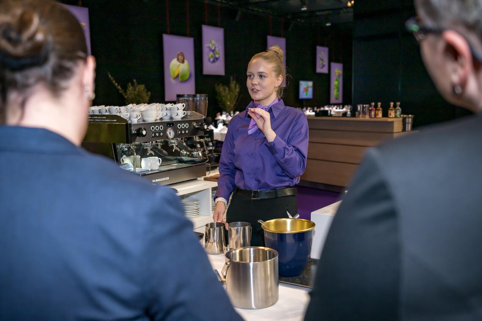 Ein Barista in einem lila Hemd, der gestikulierend zwei Personen, die ihm gegenüber am Tresen stehen, die Zubereitung von Kaffee erklärt.