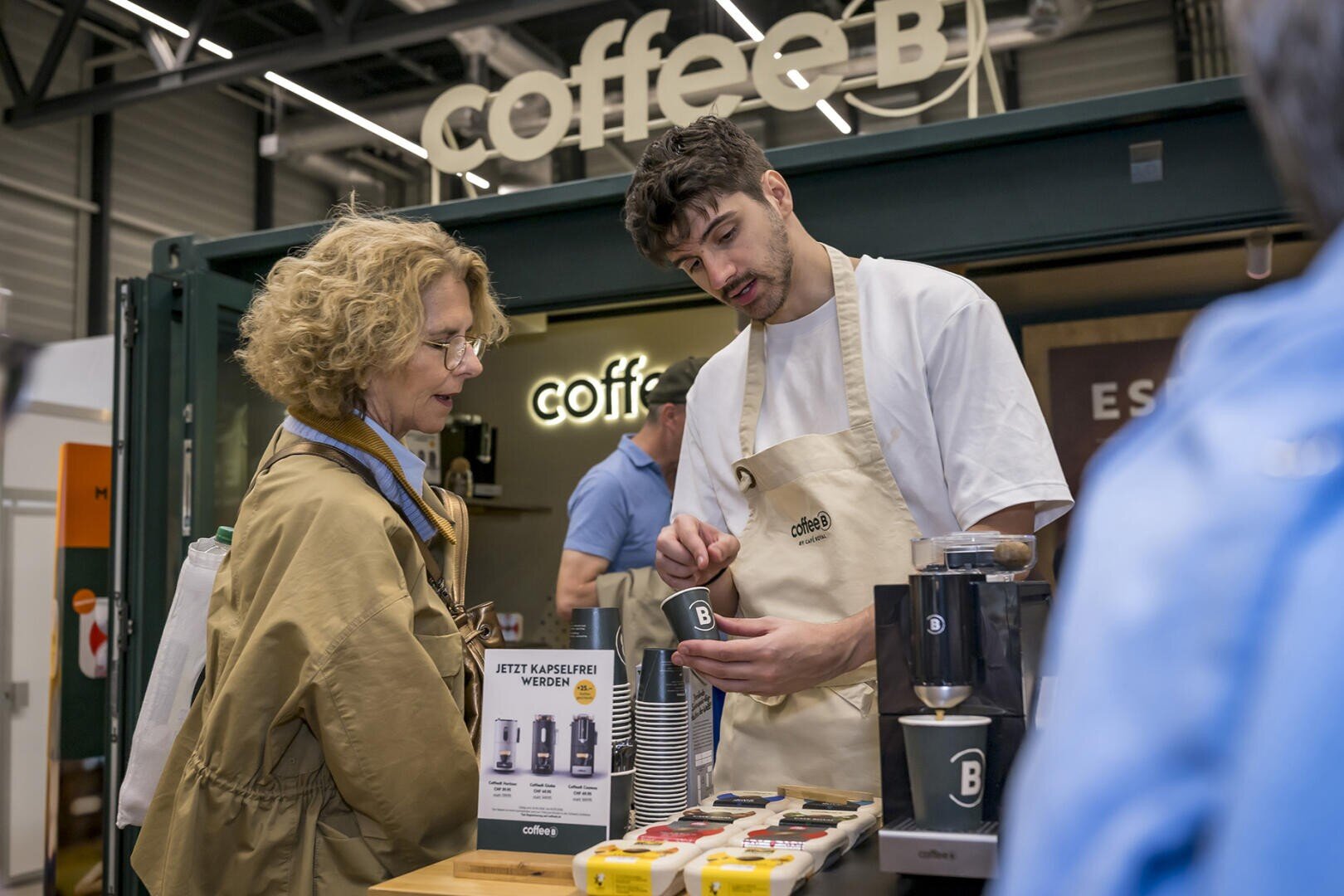 Ein junger Mann in einer Schürze präsentiert einer Frau am Stand von CoffeeB ein Kaffeeprodukt, umgeben von Kaffeemaschinen und Produktproben in einem modernen Innenraum.