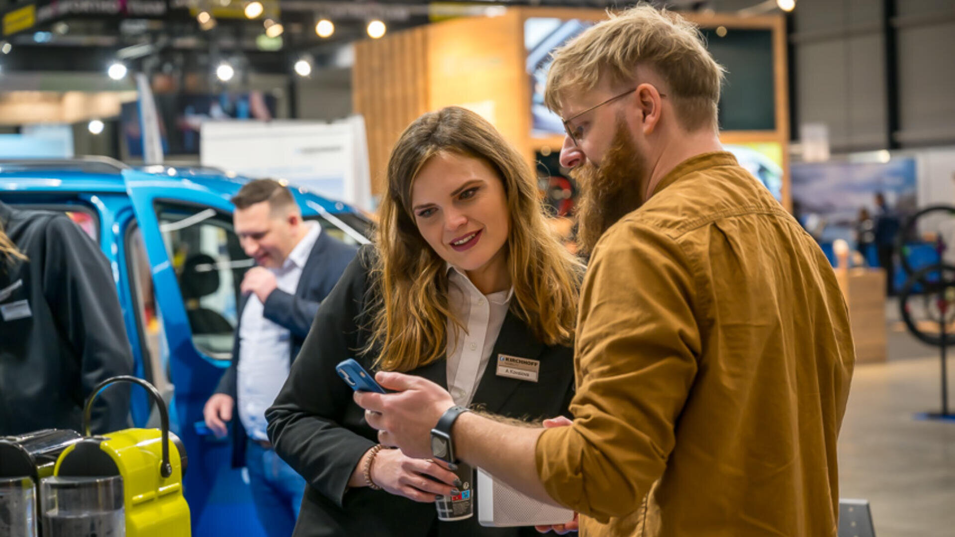Ein Mann und eine Frau stehen bei einer Indoor-Veranstaltung zusammen und schauen auf ein Smartphone. Die Frau lächelt und hält ein Getränk in der Hand. Im Hintergrund sind Menschen und ein blaues Auto zu sehen, was auf eine Messe oder Ausstellung schließen lässt.