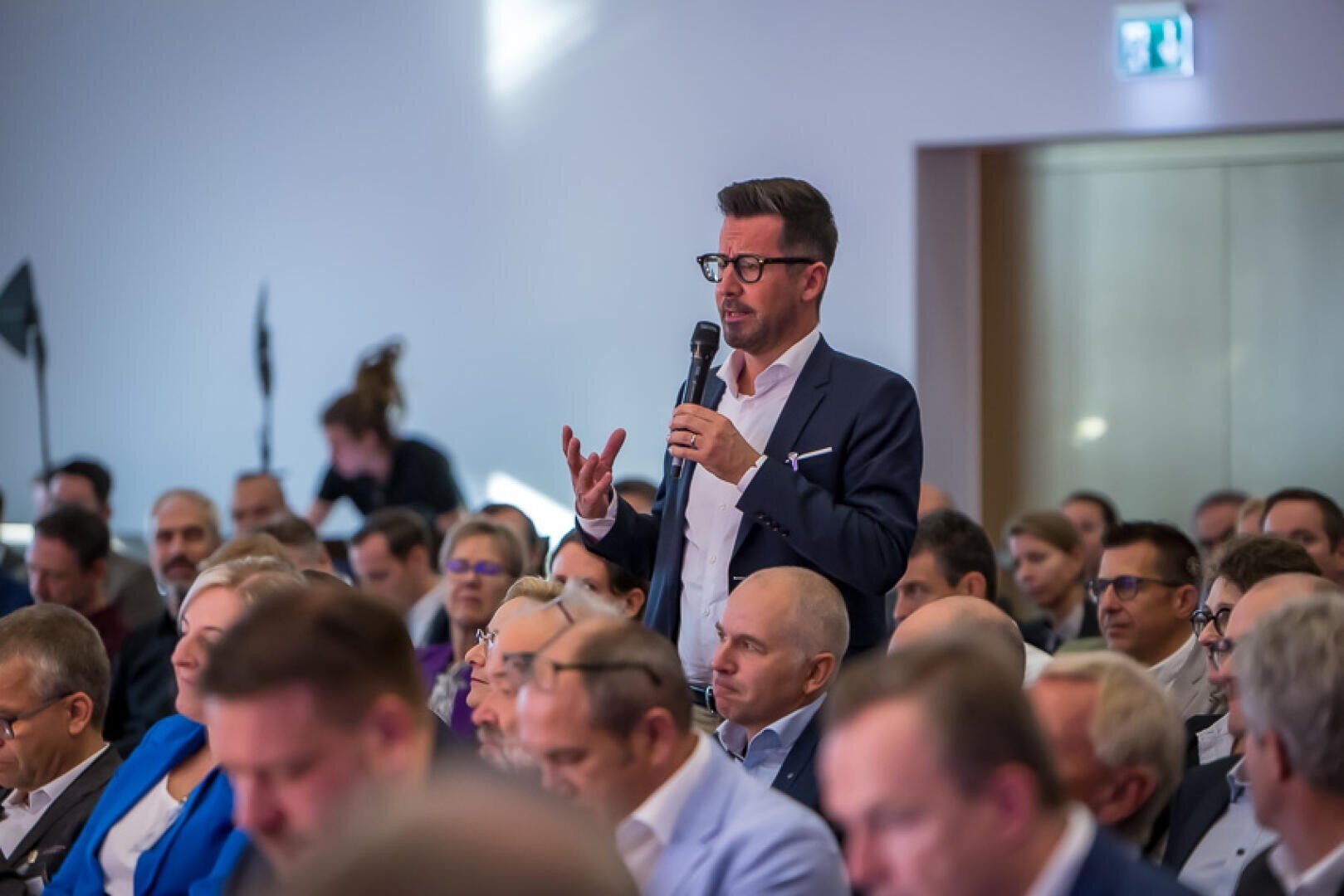Ein Mann im Anzug steht inmitten eines sitzenden Publikums, hält ein Mikrofon in der Hand und spricht, während andere bei einer Veranstaltung in einem Saal aufmerksam zuhören.