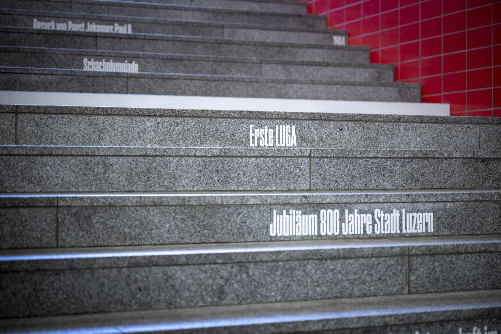 Graue Steinstufen mit aufgedrucktem deutschen Text auf den Setzstufen, darunter „Erste UGCA“ und „Jubiläum 800 Jahre Stadt Luzern“. Auf der rechten Seite ist eine rot geflieste Wand sichtbar.