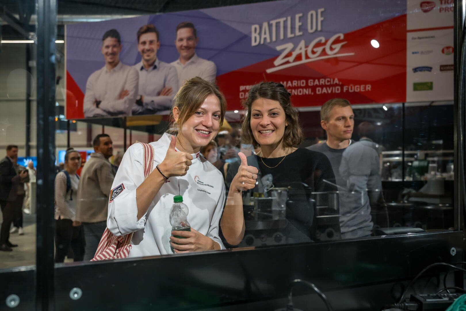 Zwei Frauen, die vor einem Werbebanner zur Battle of Zagg den Daumen hochhalten.