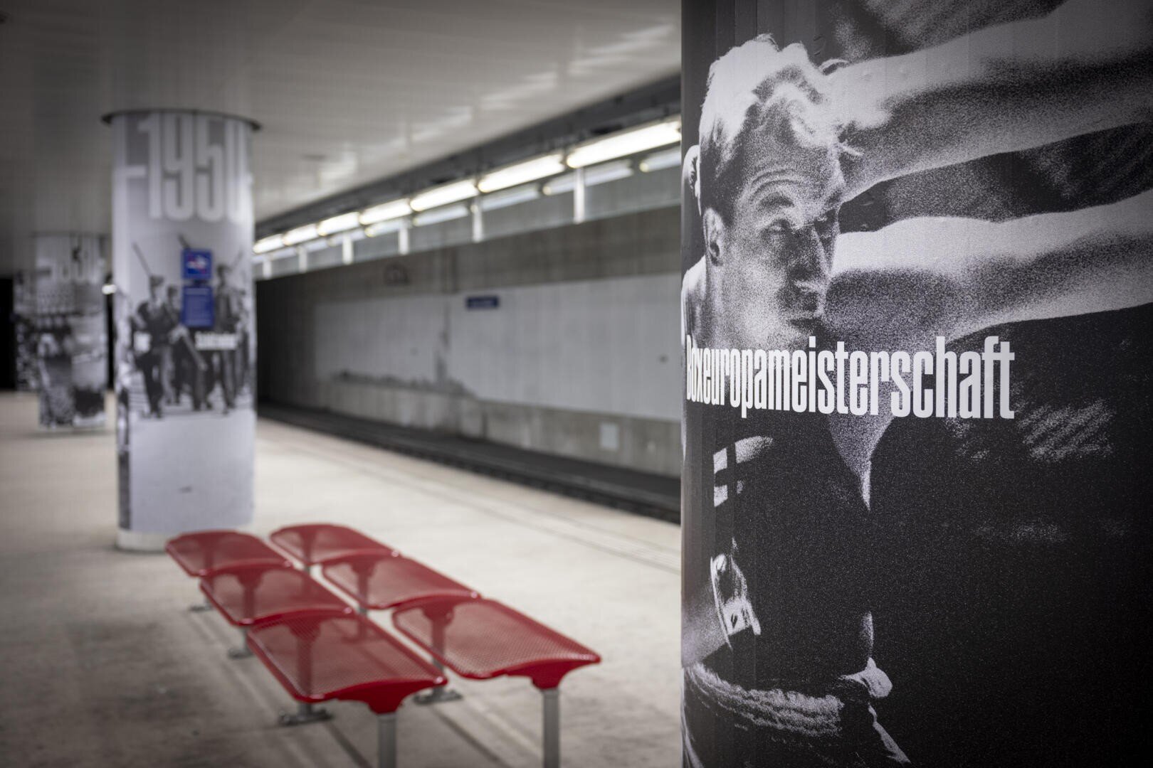 Ein U-Bahnsteig mit roten Bänken und Säulen, die mit schwarz-weißen Sportbildern umhüllt sind. Eine Säule zeigt einen Boxer und das Wort „Europameisterschaft“. Der Bahnsteig ist leer und modern.