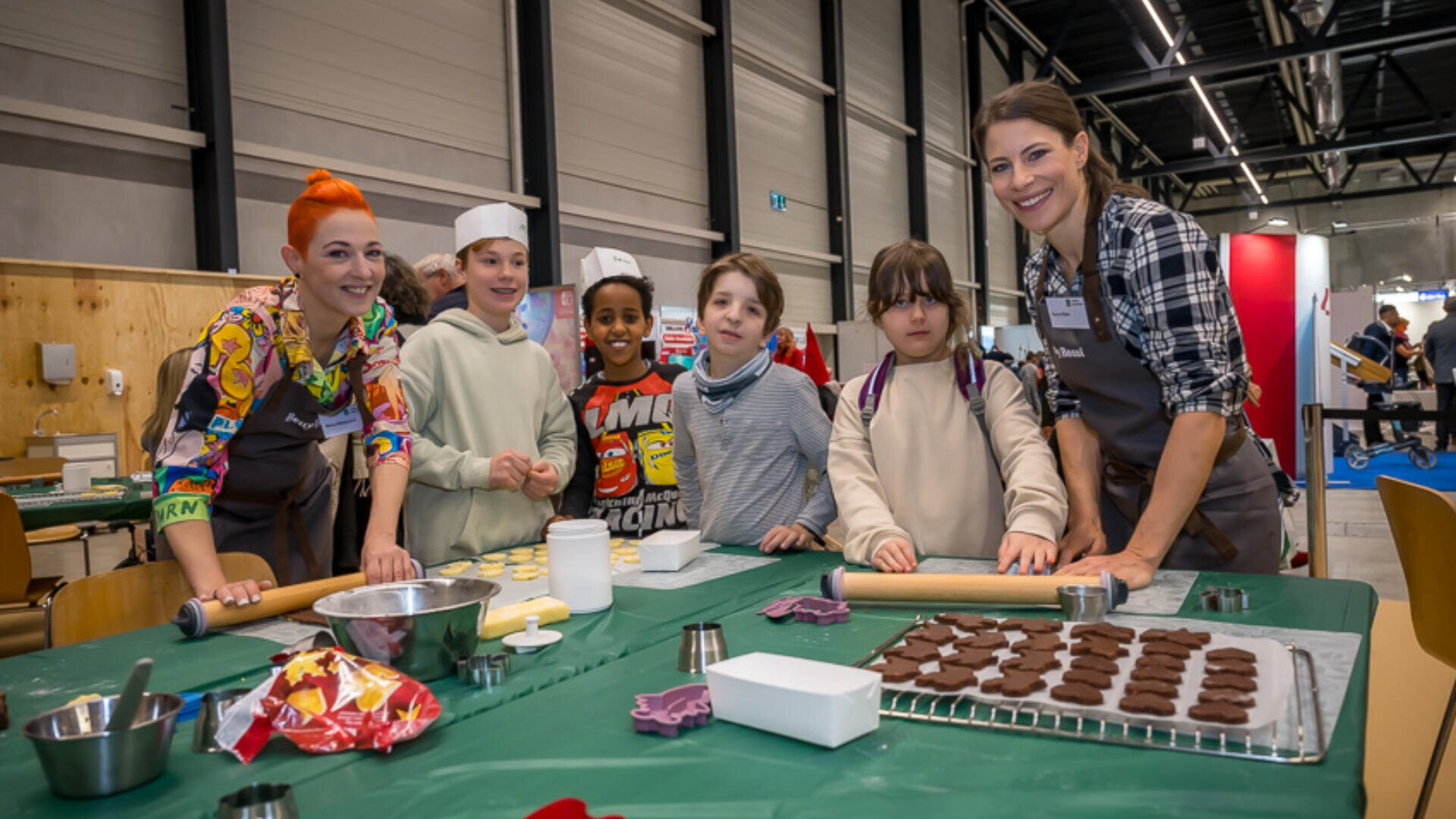 Eine Gruppe von Kindern und zwei Erwachsenen steht lächelnd um einen Tisch und backt gemeinsam Plätzchen. Ausstechformen, Teig und Backzubehör liegen in einem hellen Innenraum auf dem Tisch ausgebreitet.