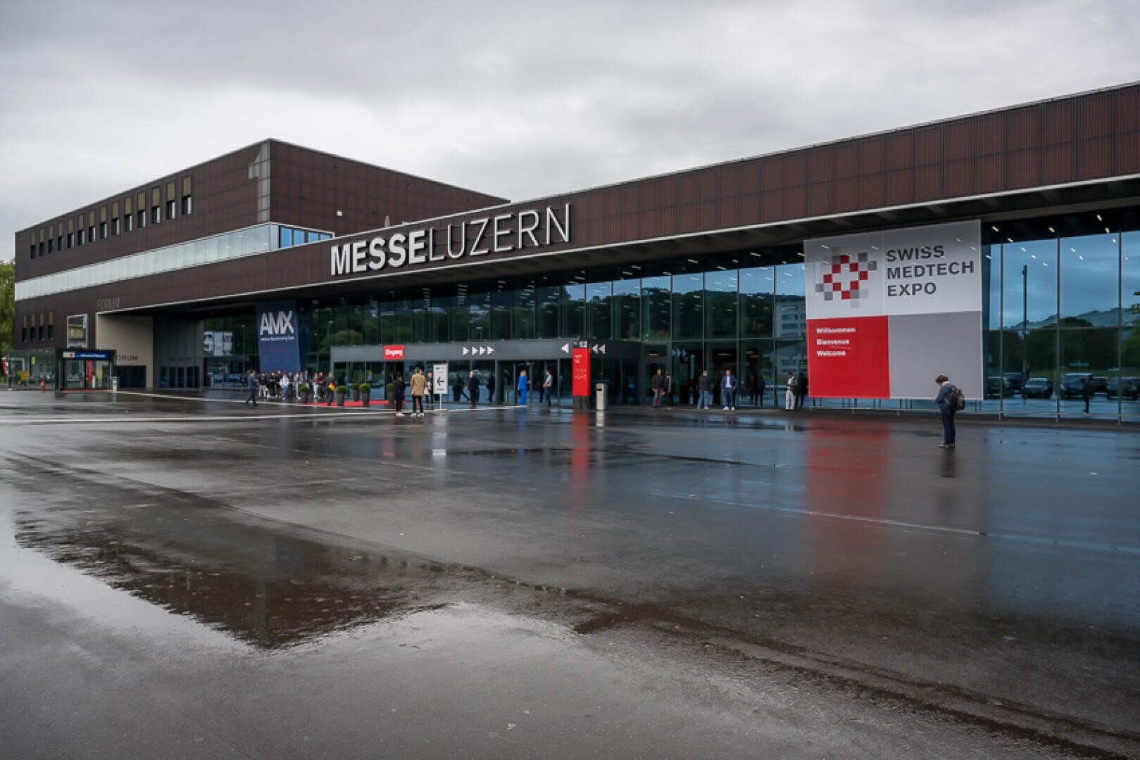 Das Äussere des Kongresszentrums der Messe Luzern an einem bewölkten Tag, mit der Beschilderung der Swiss Medtech Expo und den Menschen, die das Gebäude betreten. Das nasse Pflaster reflektiert die Struktur und den Eingangsbereich.