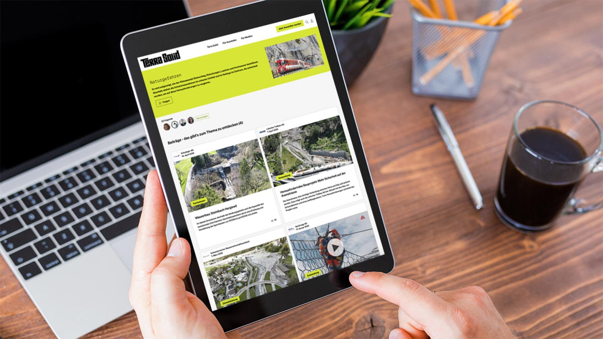 Eine Person hält ein Tablet, auf dem eine Website mit Luftbildern und Artikeln angezeigt wird. Die Szene umfasst einen Laptop, einen Notizblock mit Bleistift, eine Brille und eine Tasse Kaffee auf einem Holztisch.