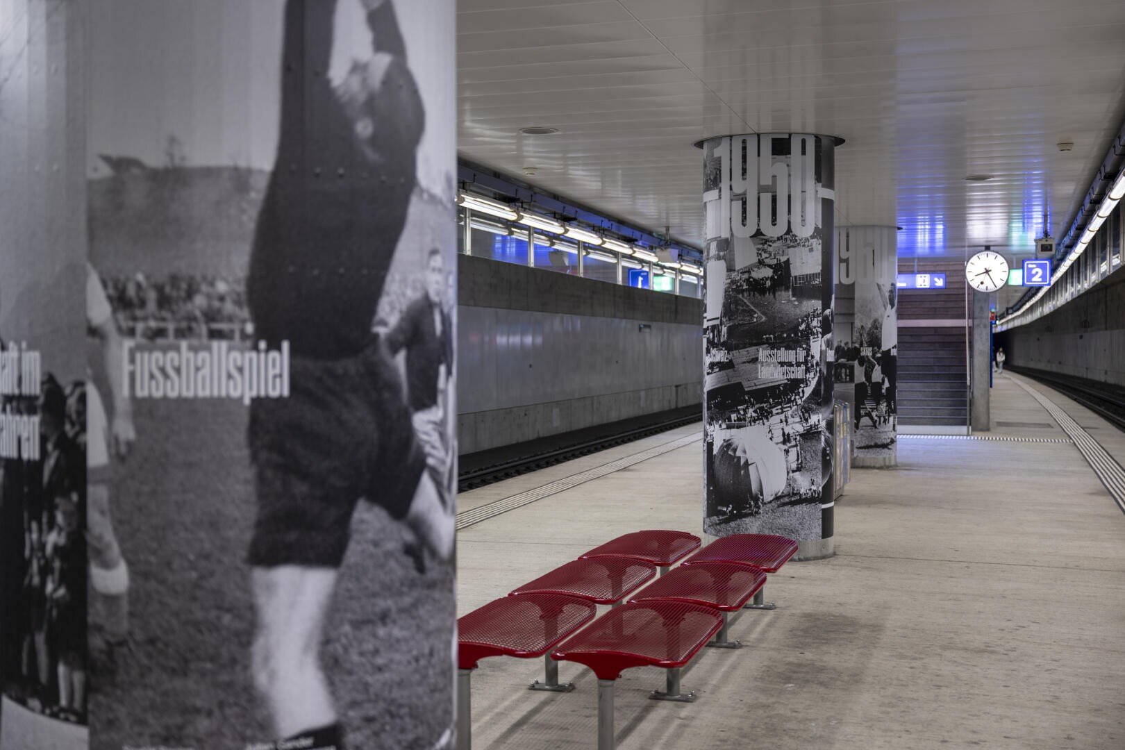 U-Bahnsteig mit roten Bänken, Säulen mit schwarz-weißen Sportfotos und dem Wort „Fußballspiel“, einer Uhr, die 10:01 zeigt, und im Hintergrund eine Treppe, die nach oben führt.